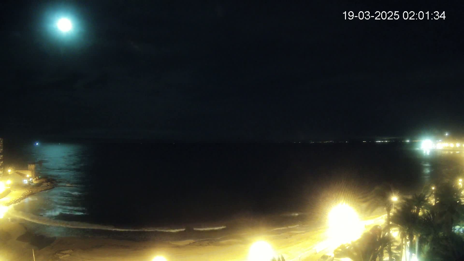 A nighttime view of a beach with gentle waves under a bright moon and partly cloudy sky, illuminated by streetlights along the shore.