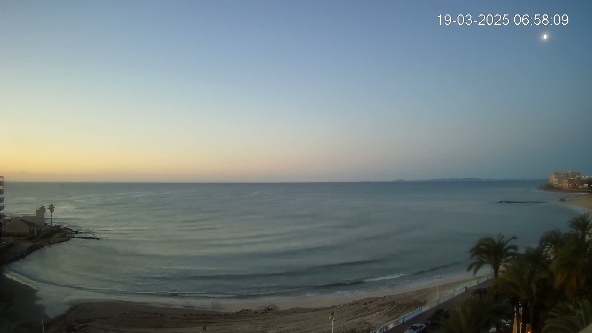 A calm, pre-dawn ocean scene shows a sandy beach, palm trees, buildings, and a mostly clear sky with a hint of sunrise.
