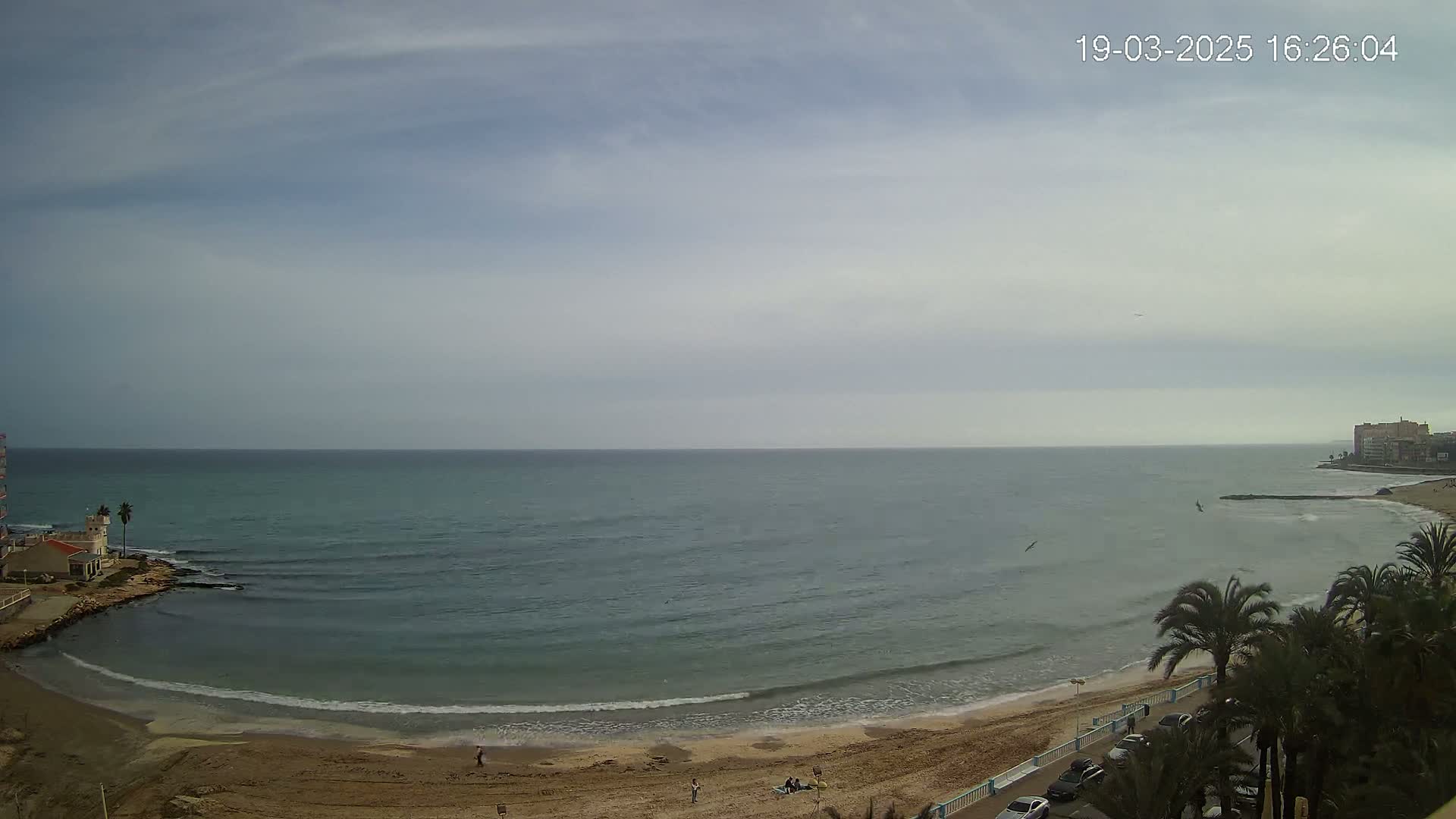A sandy beach curves around a calm, teal ocean under a mostly cloudy sky.