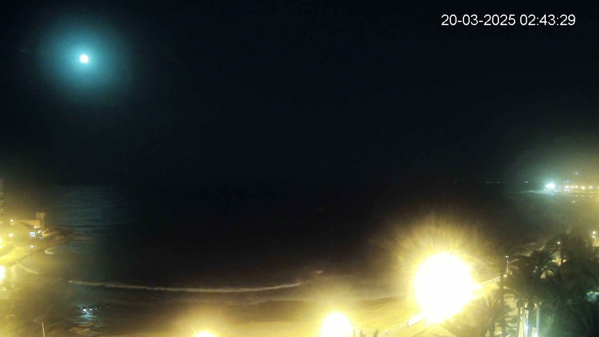 A bright moon illuminates a dark, nighttime beach scene with gentle waves and illuminated palm trees along a shoreline.