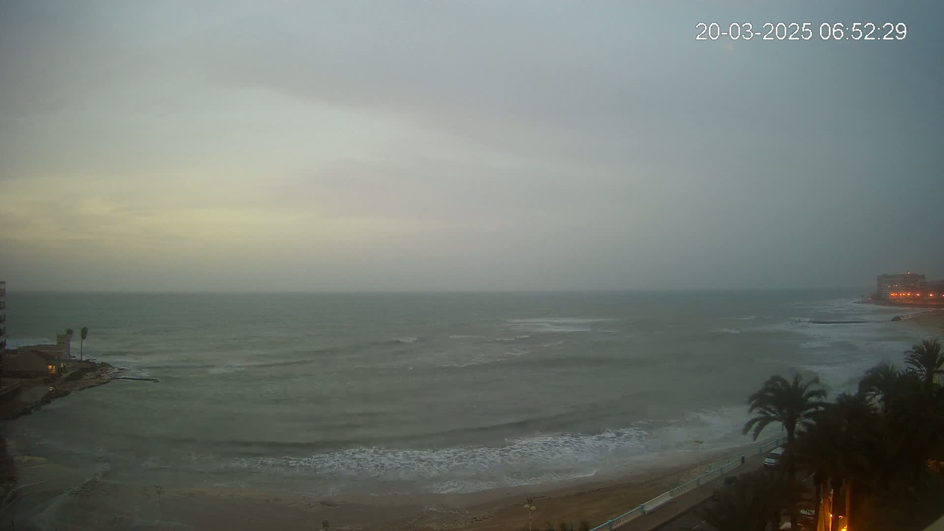 A mostly cloudy, overcast day shows a choppy sea with small waves lapping a sandy beach, palm trees, and buildings visible in the distance.
