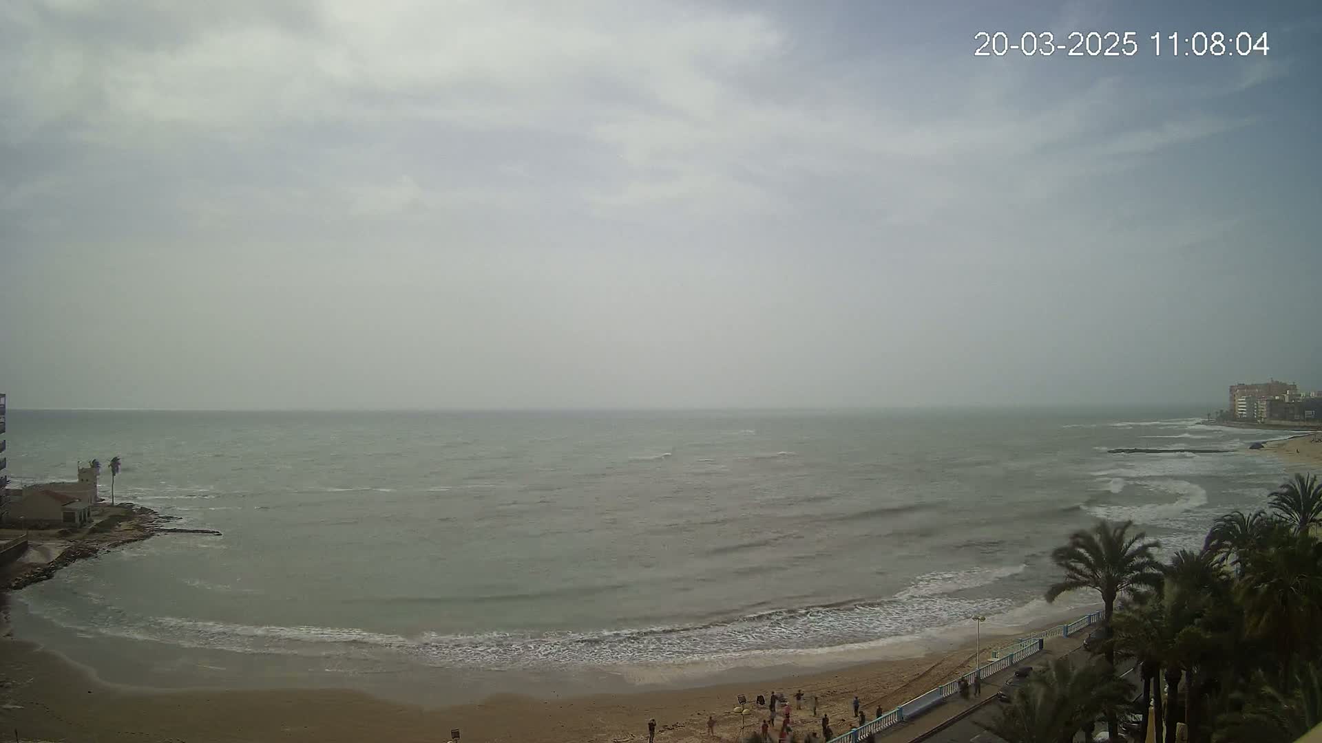 A hazy, overcast day shows a beach with a few people, palm trees, and a calm sea with small waves.