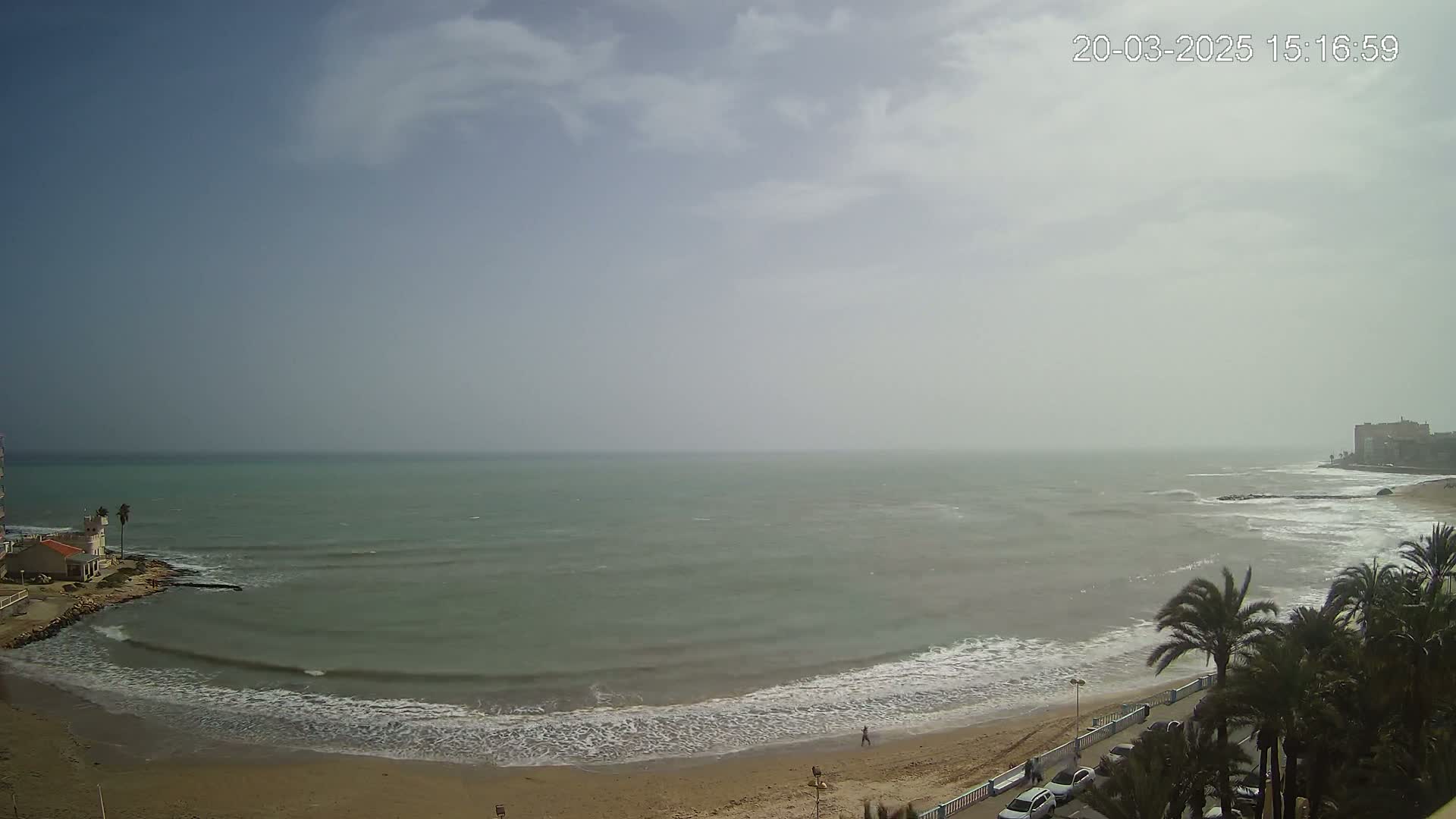 A hazy, overcast day shows a sandy beach with small waves lapping the shore, palm trees lining a nearby road, and a calm sea extending to a distant coastline.