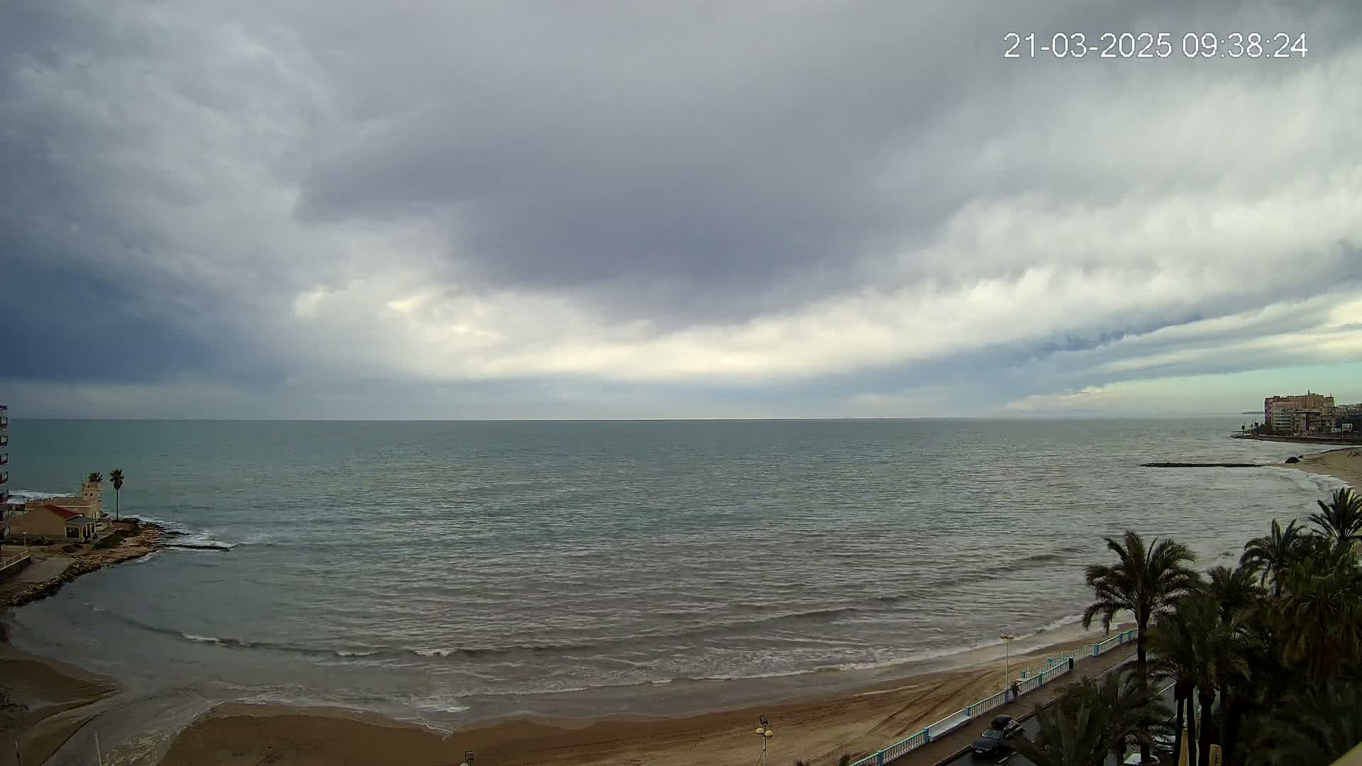 A mostly cloudy sky hangs over a calm sea with a sandy beach and palm trees lining a promenade.