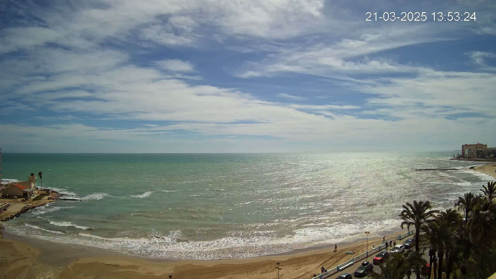A sandy beach with gently breaking waves stretches along a calm, greenish-blue sea under a partly cloudy sky.