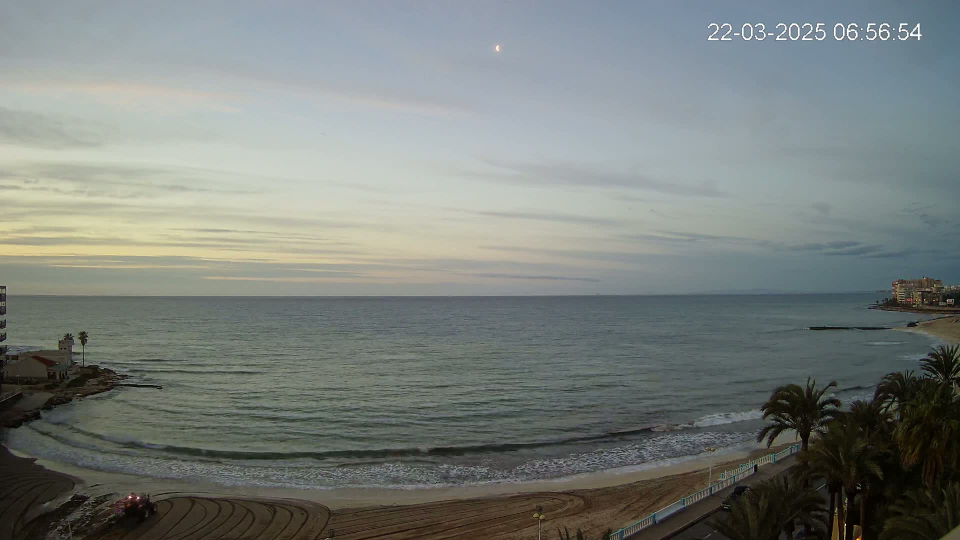 A crescent moon hangs in the twilight sky over a calm sea lapping a sandy beach lined with palm trees.