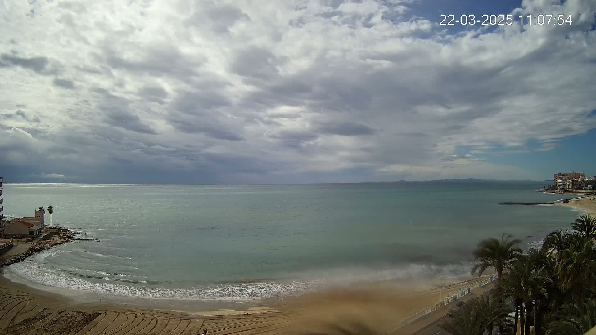 A mostly cloudy day shows a calm ocean meeting a sandy beach lined with palm trees and buildings in the distance.
