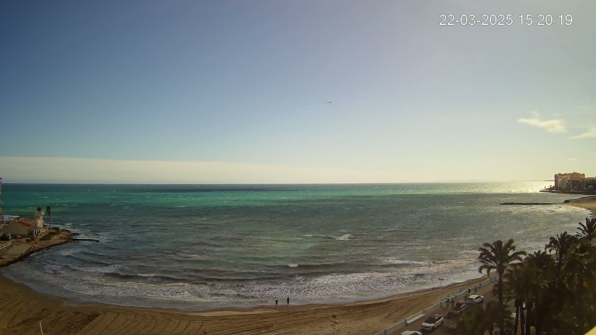A sandy beach with palm trees and buildings is visible next to a wavy, teal ocean under a mostly clear sky.