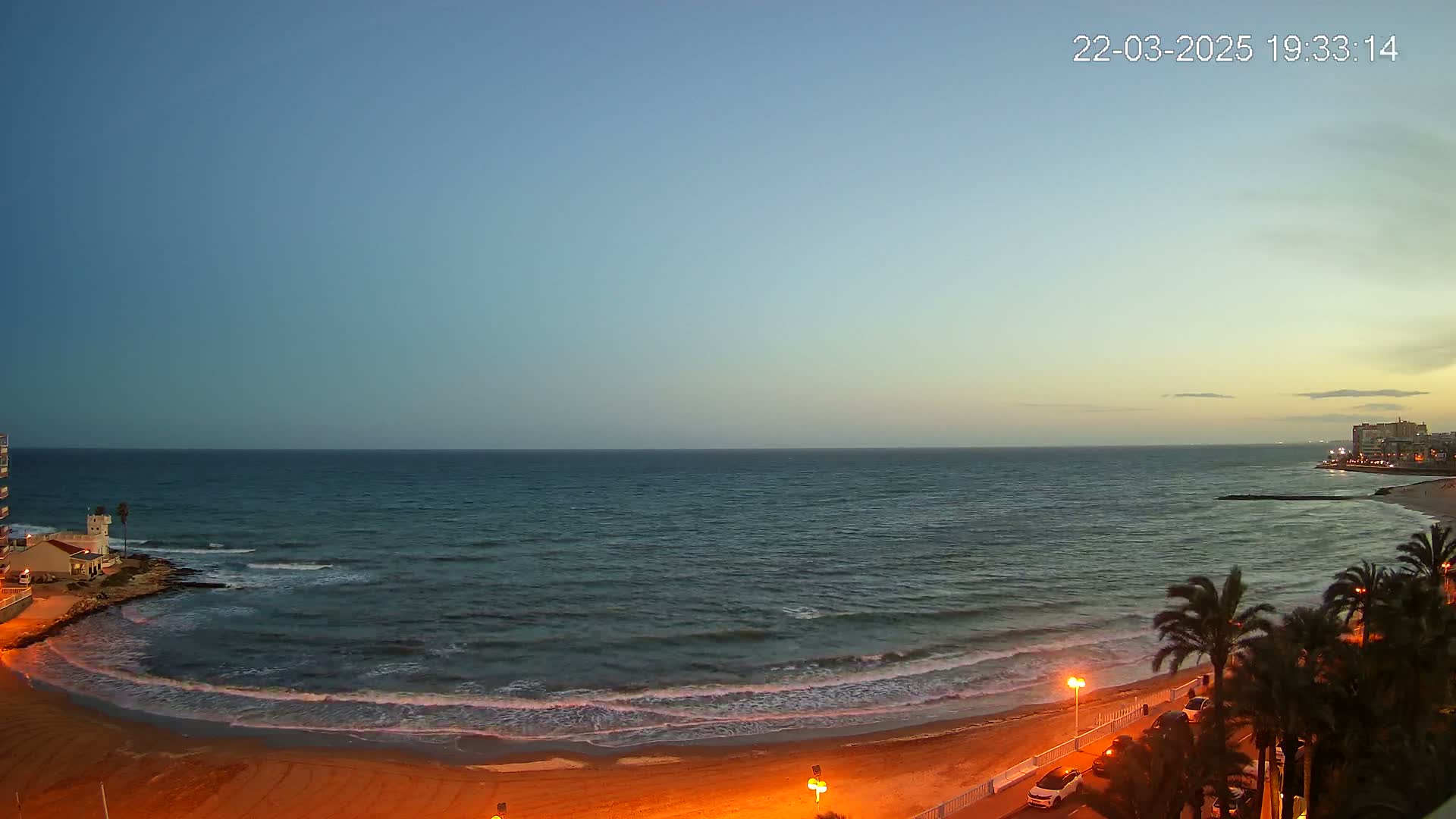 A sandy beach at dusk with gentle waves lapping the shore, palm trees lining a nearby road, and buildings visible in the distance.