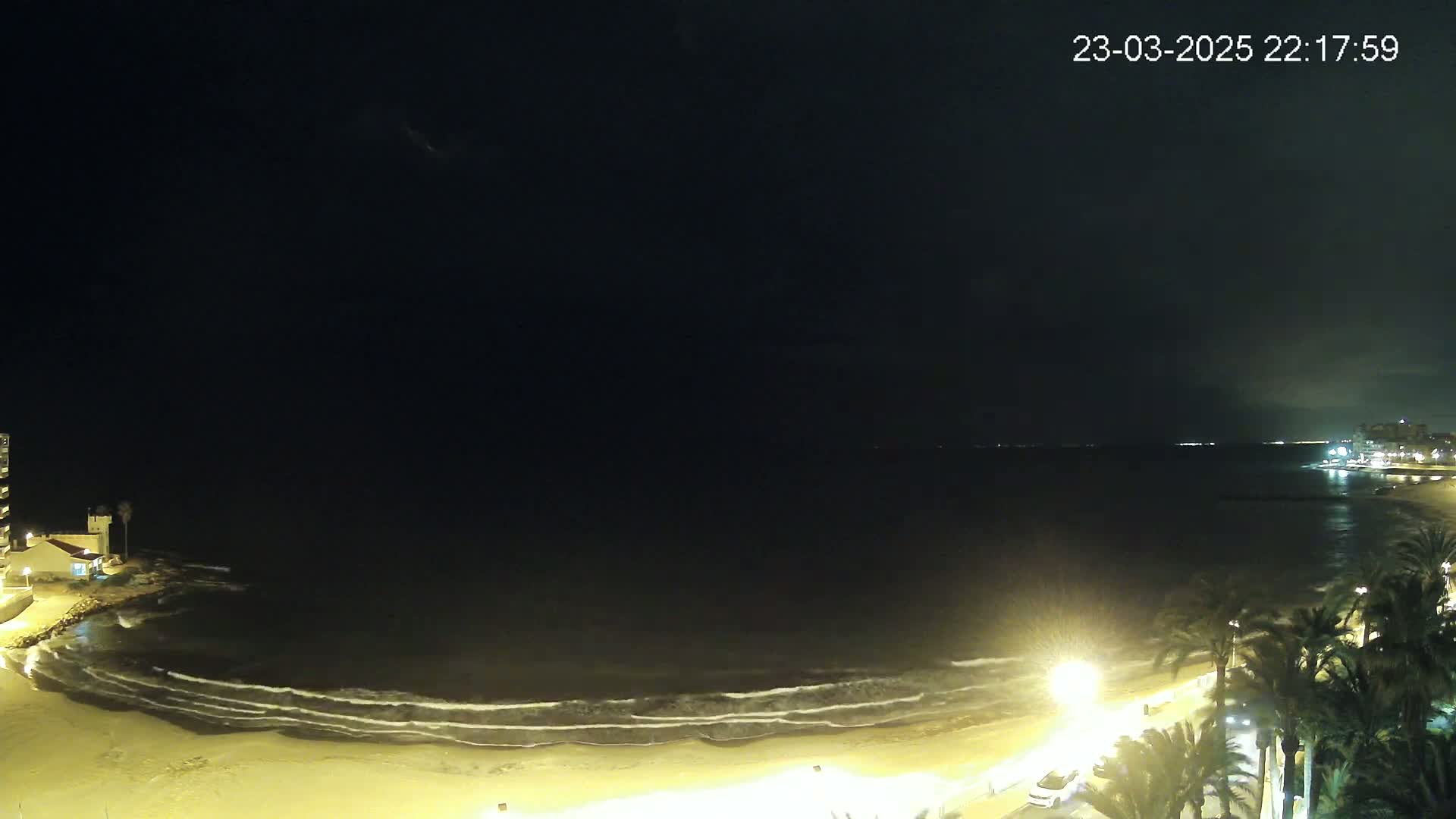 A nighttime beach scene shows gently lapping waves on a sandy shore, palm trees lining a dimly lit promenade, and a building in the distance under a dark, clear sky.