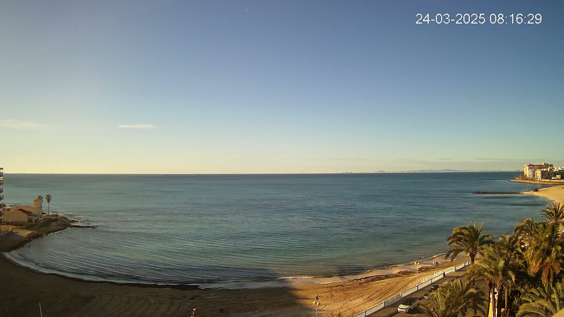 A calm, clear day reveals a sandy beach, palm trees, buildings, and a tranquil sea stretching to a distant shoreline.