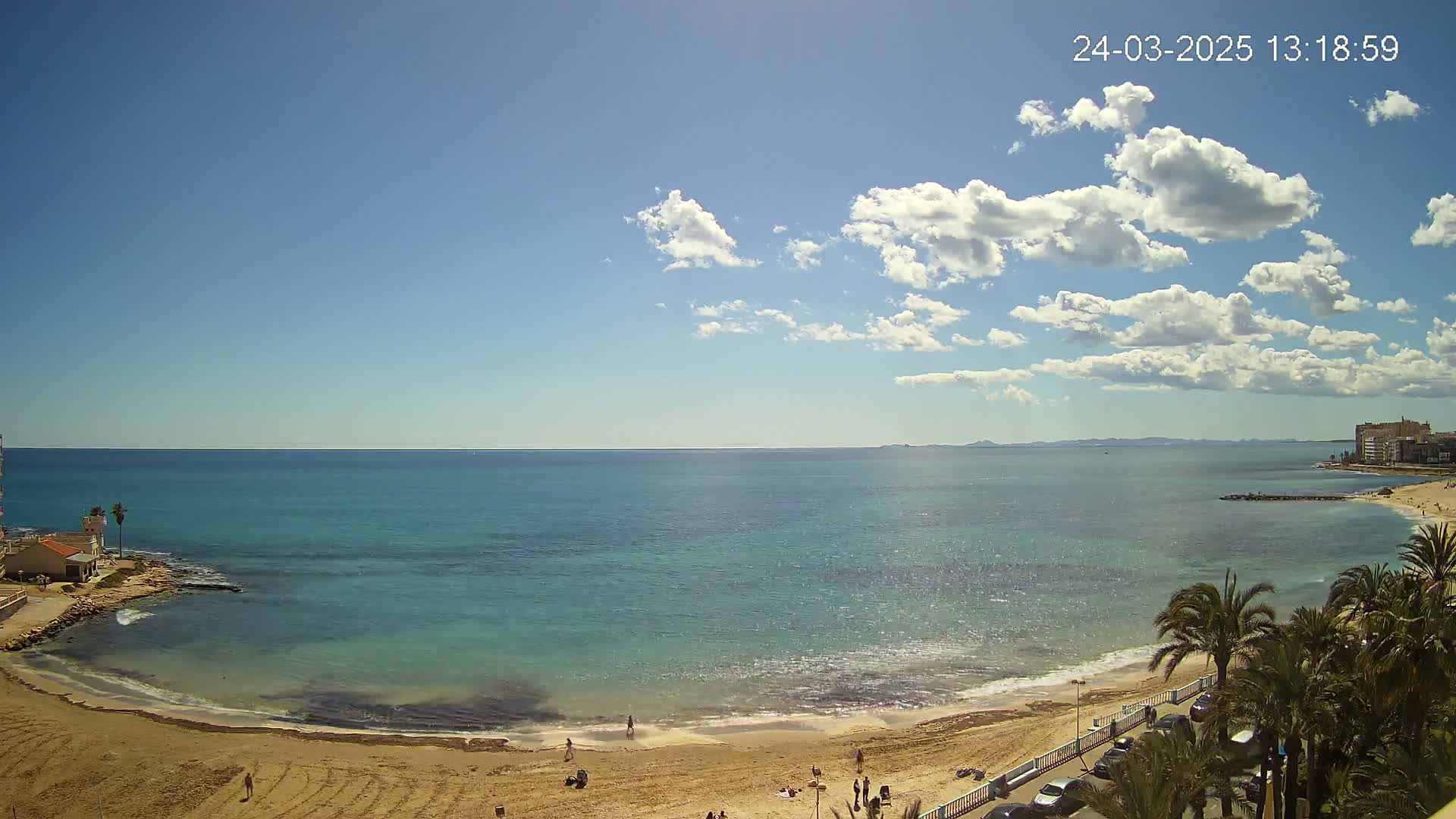 A sandy beach curves along a calm, turquoise sea under a partly cloudy blue sky.