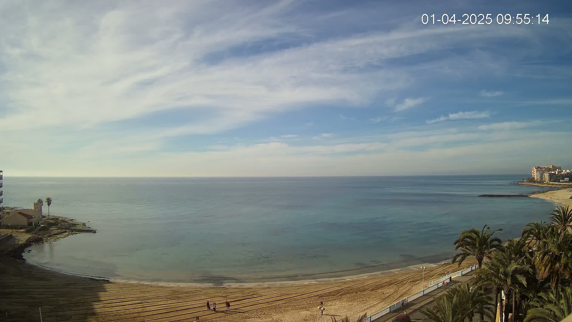 A sandy beach curves around a calm, teal ocean under a mostly sunny sky with scattered clouds.