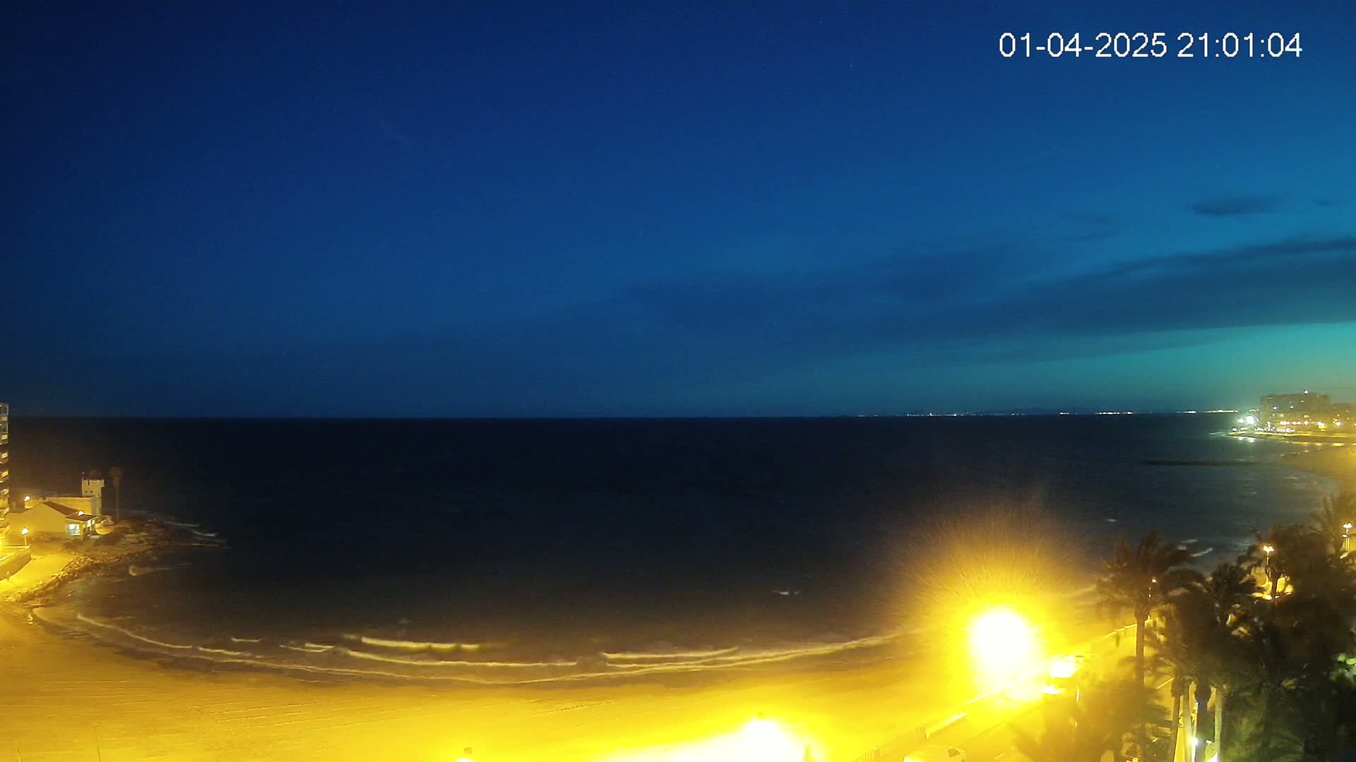 A nighttime view of a beach with gentle waves lapping the shore under a dark blue, mostly clear sky.