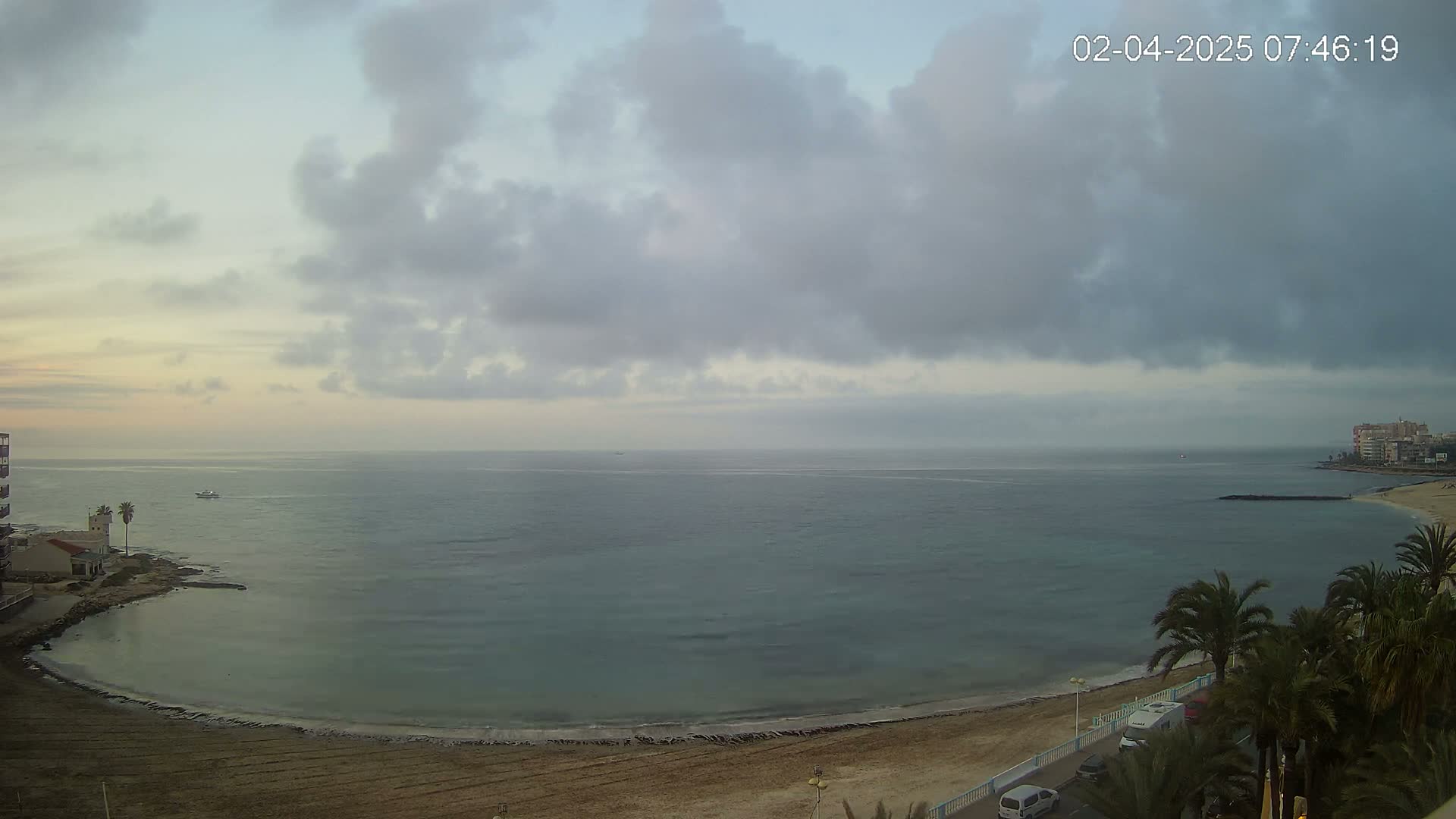 A mostly cloudy morning scene shows a calm sea curving around a sandy beach with palm trees and buildings in the background.