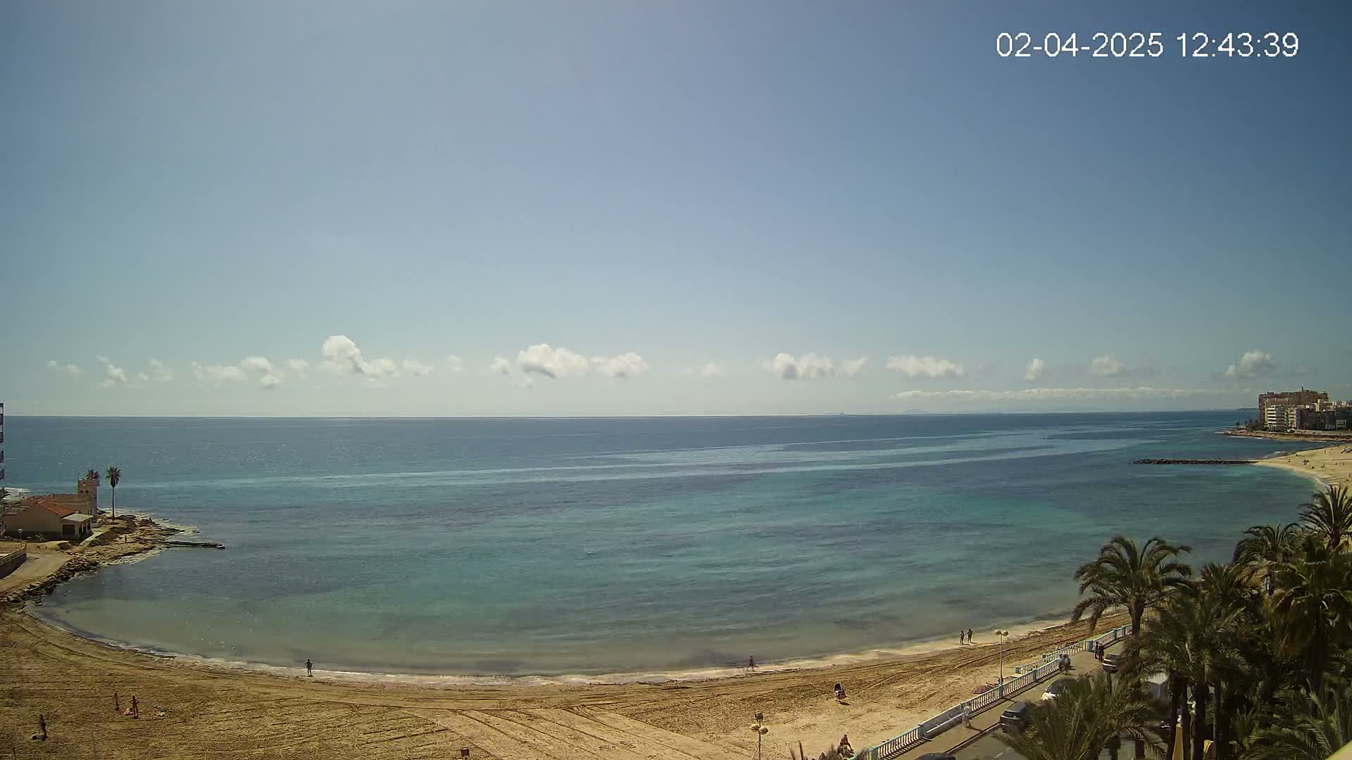 A sandy beach curves around a calm, teal-colored sea under a mostly sunny sky with a few scattered clouds.
