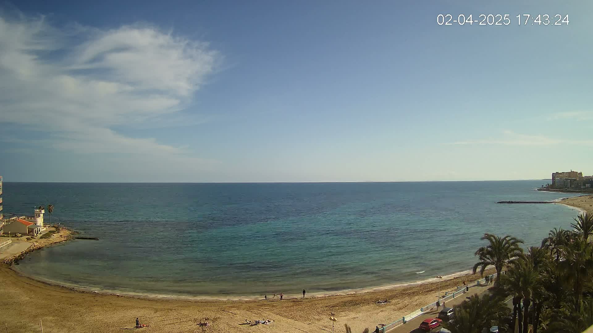 A sandy beach curves around a calm, teal sea under a mostly clear sky with a few scattered clouds.