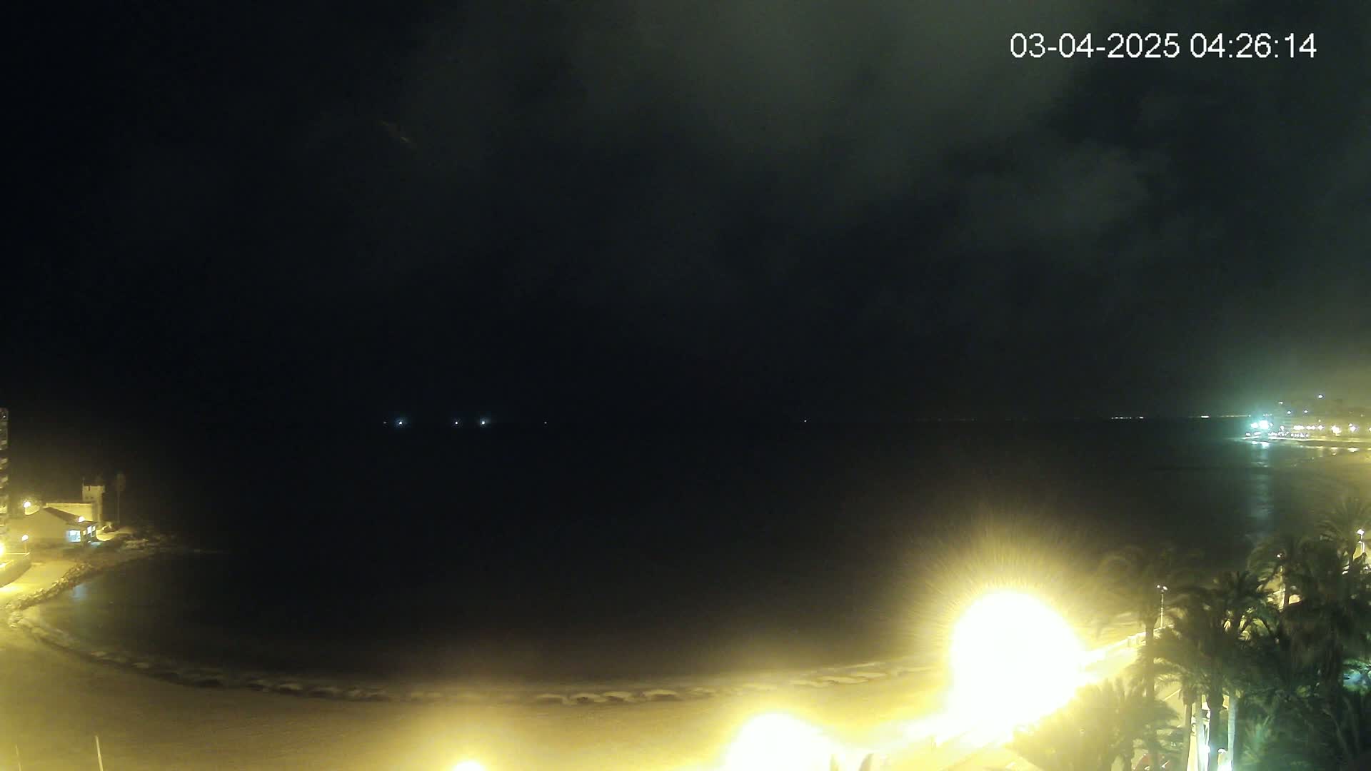 A nighttime view of a dark ocean bay, illuminated by lights on a beach with palm trees.