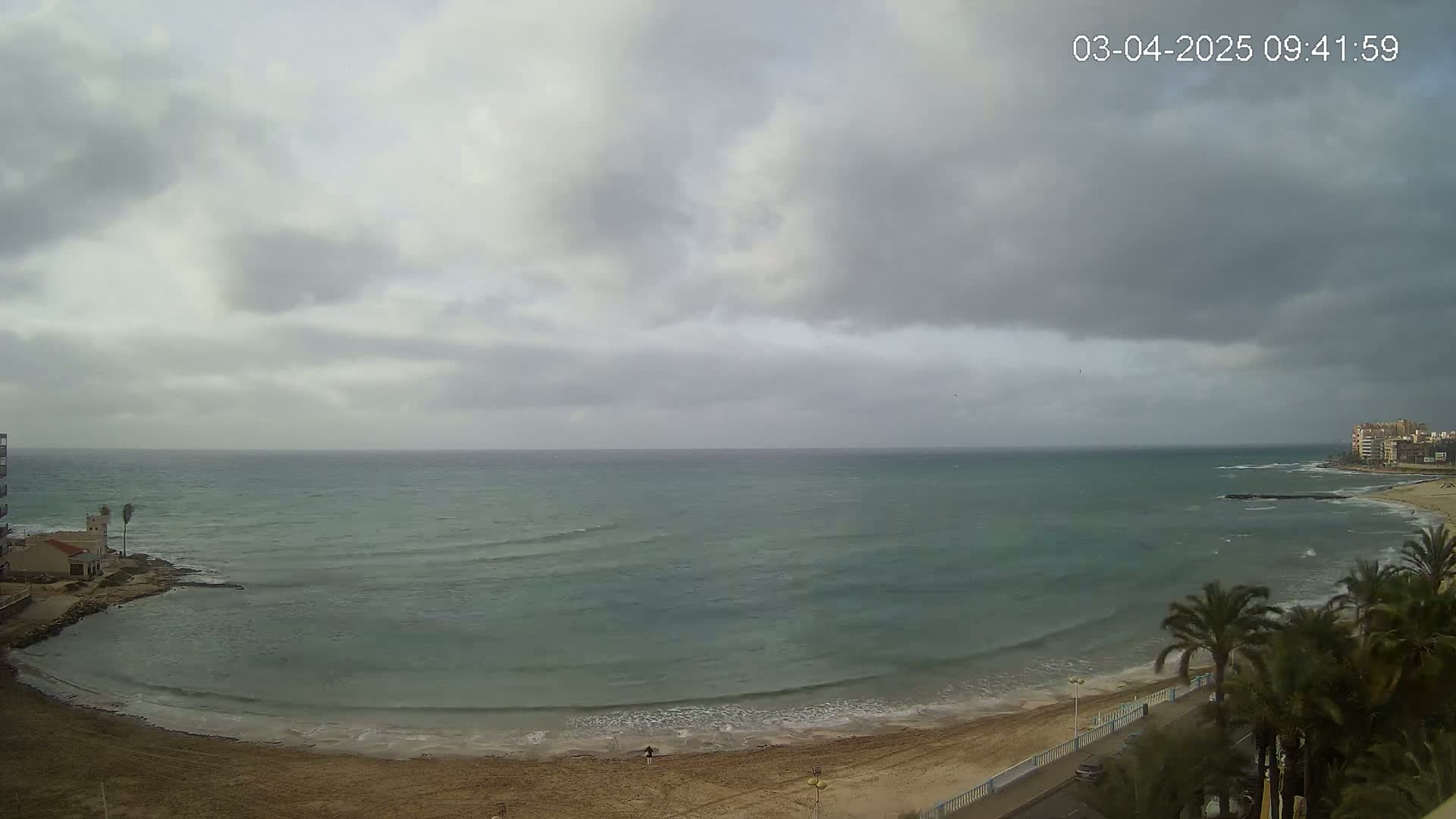 A mostly cloudy day overlooks a calm sea with small waves lapping a sandy beach lined with palm trees and buildings.