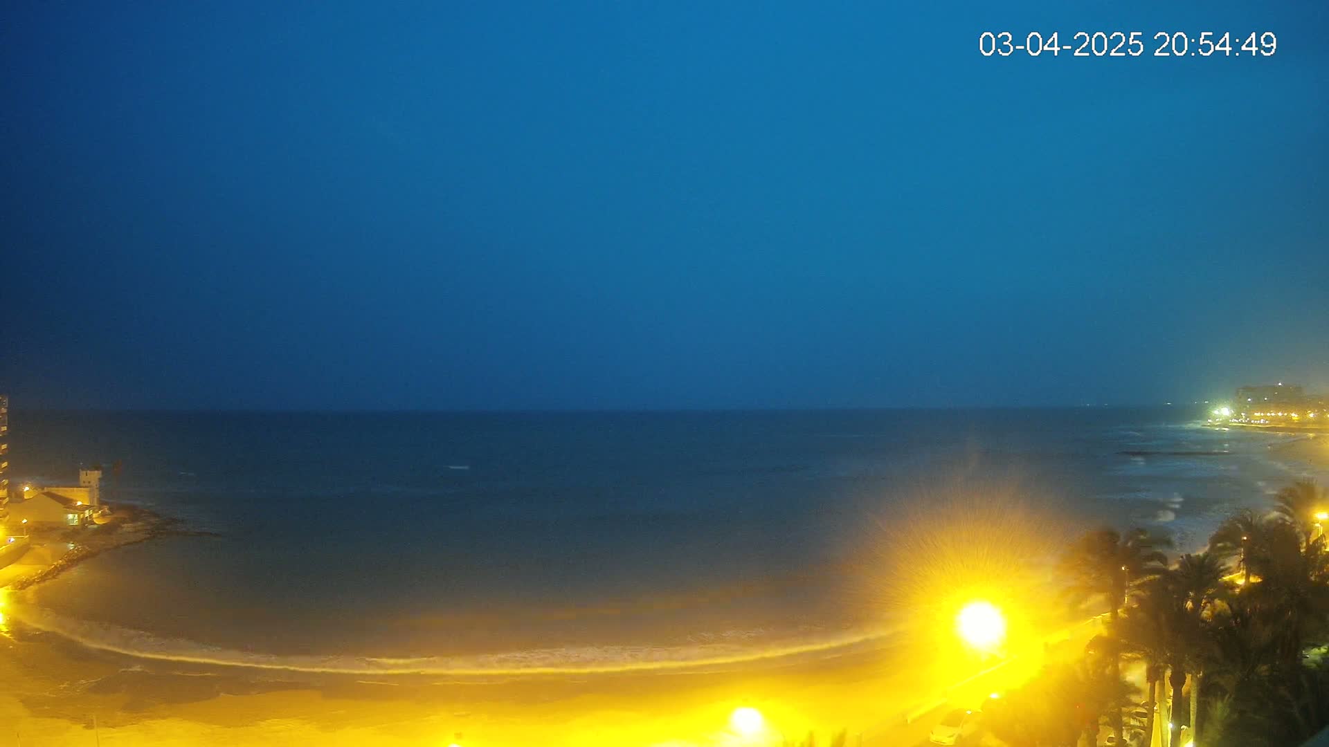 A nighttime beach scene shows a dark blue sky, a calm ocean, and a sandy shore illuminated by artificial lights.