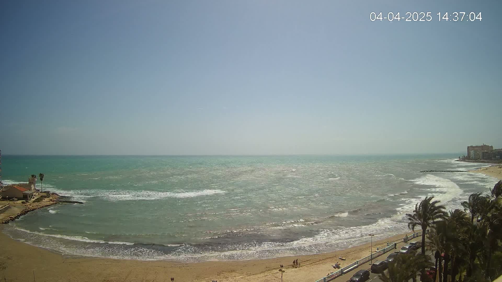 A sandy beach with palm trees and buildings along the shore meets a somewhat choppy, teal-colored sea under a clear, sunny sky.