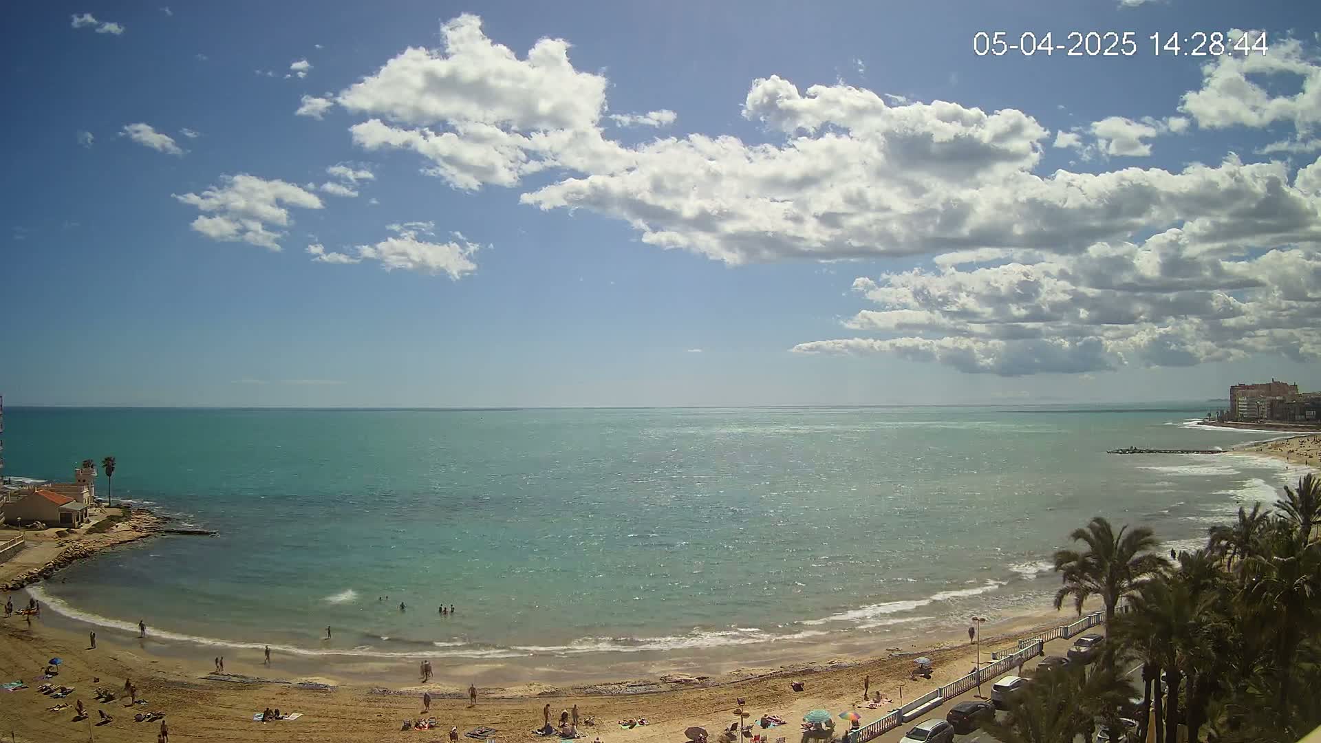 A sandy beach with scattered people is visible, bordered by palm trees and buildings, next to a calm, teal-colored sea under a partly cloudy sky.