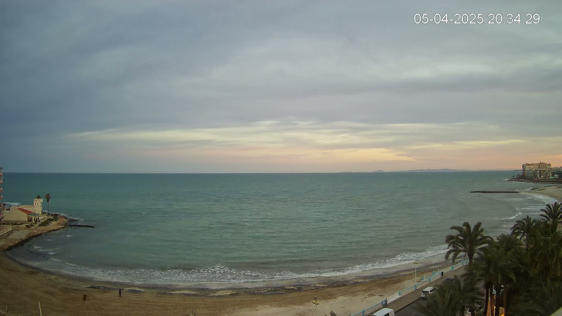 A sandy beach curves along a calm, greenish-grey sea under a mostly cloudy, pale-pink sunset sky.