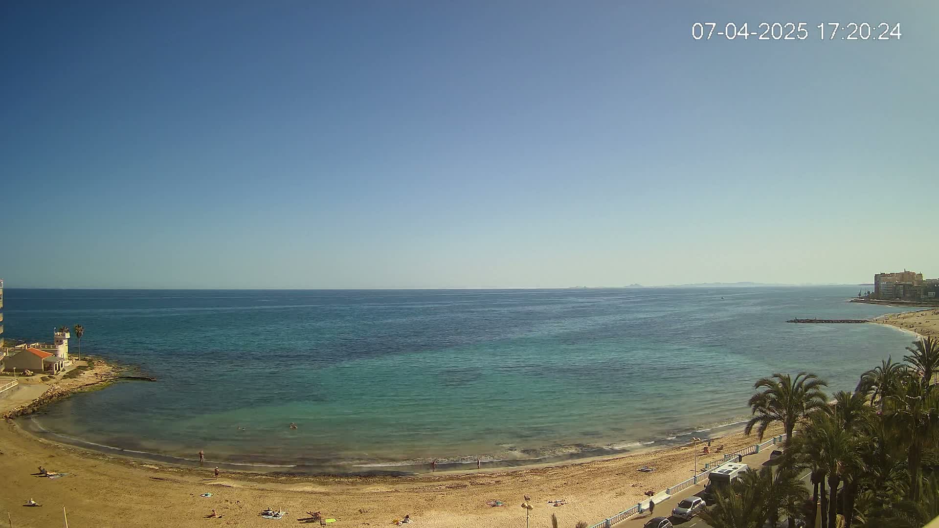 A sandy beach curves around a calm, clear turquoise sea under a bright, sunny sky.