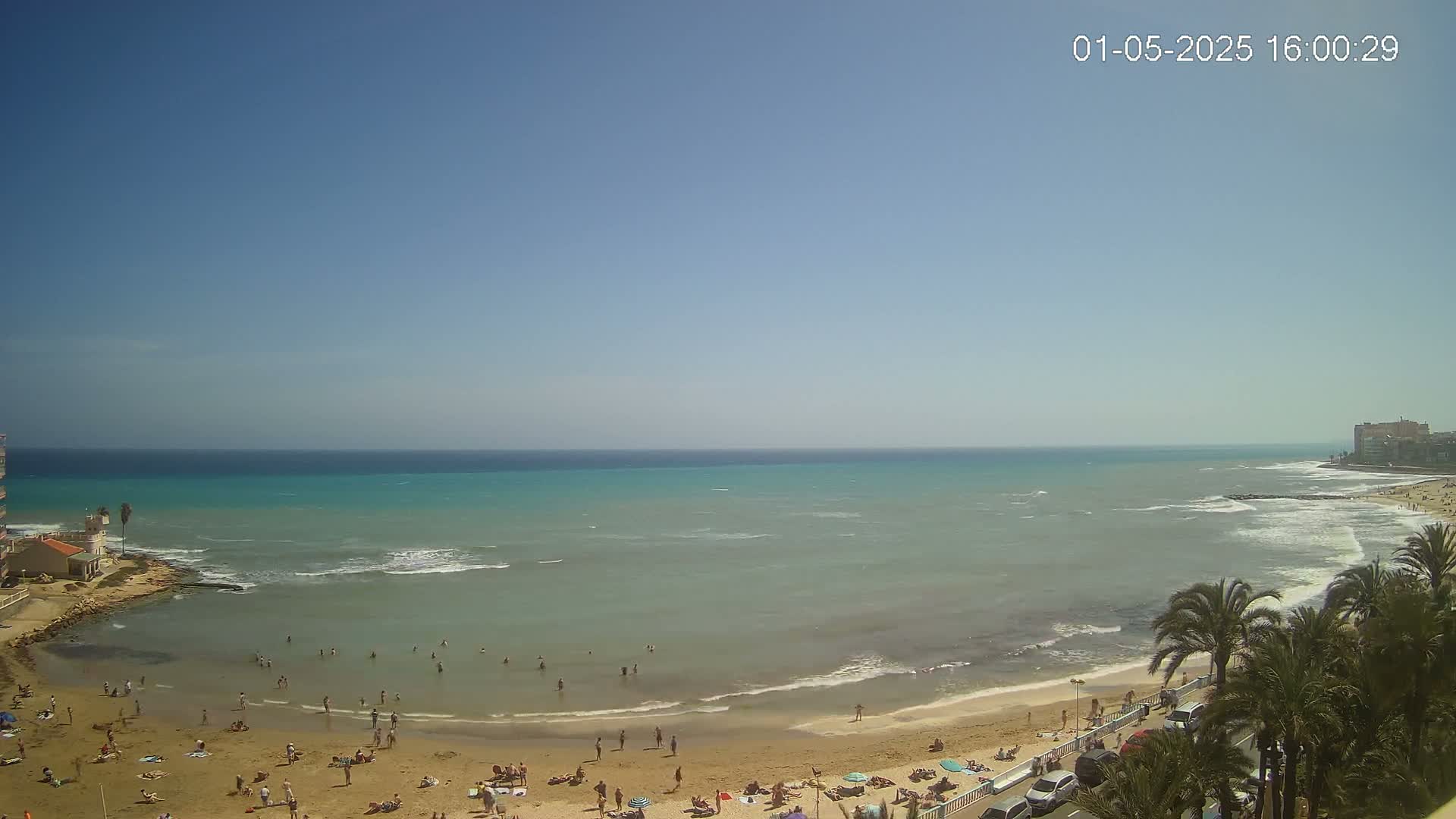 A sandy beach with scattered sunbathers is visible next to a calm, light-green ocean under a clear blue sky.