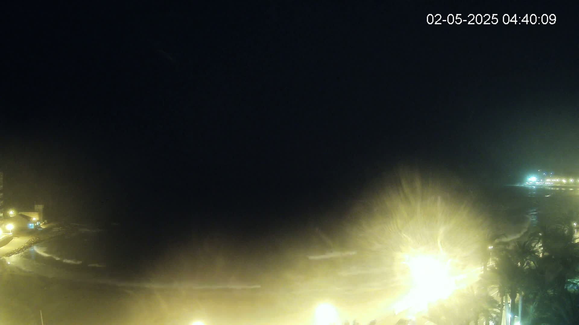 A nighttime coastal scene shows tumultuous waves crashing against a shoreline illuminated by streetlights under a dark, stormy sky.