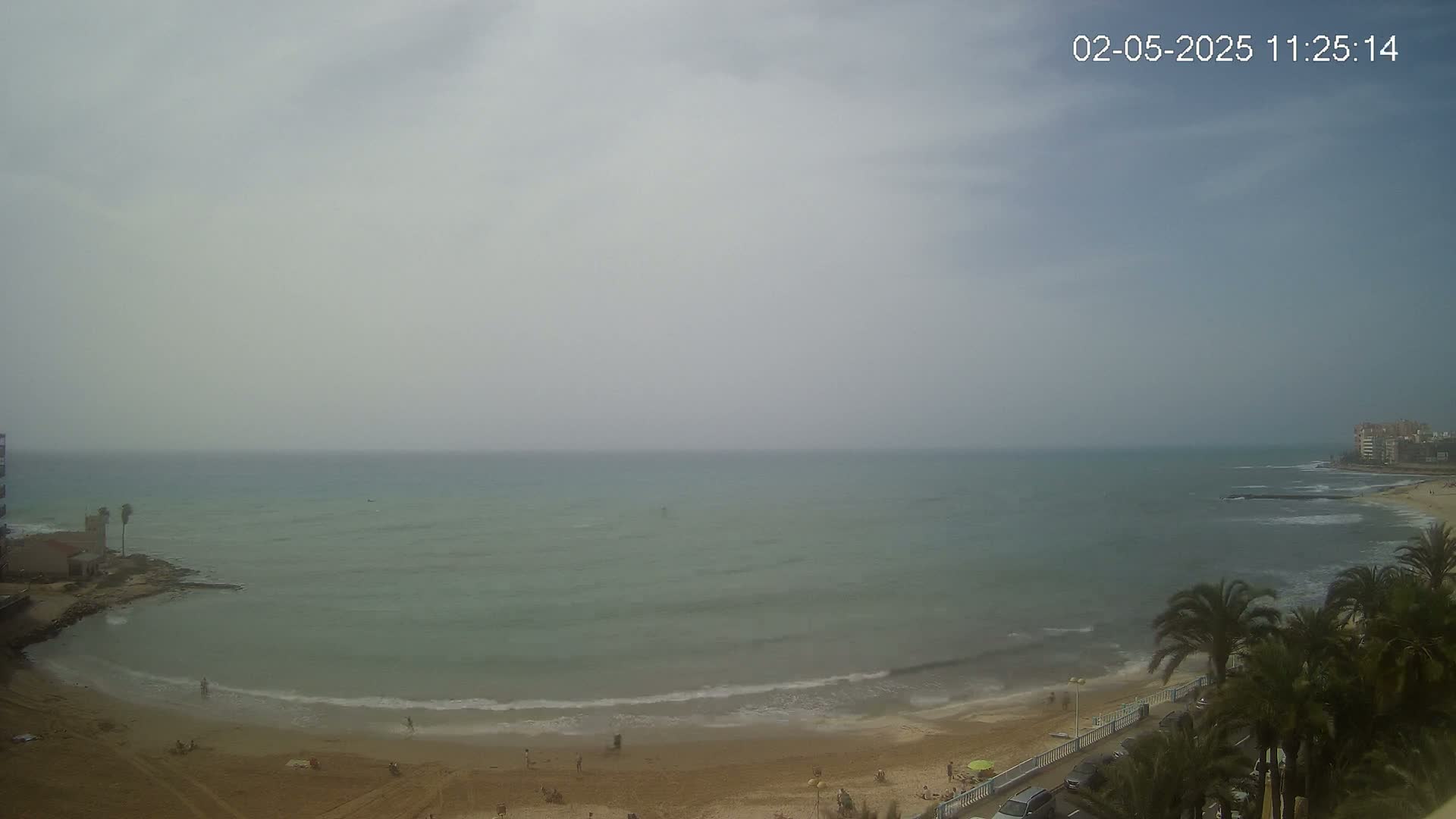 A hazy, overcast day reveals a sandy beach with a few people, gentle waves lapping the shore, and palm trees lining a nearby road overlooking a calm sea.