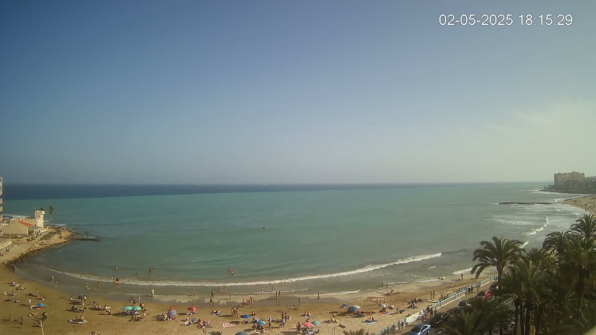 A sandy beach with people sunbathing and swimming is bordered by calm, turquoise water under a clear blue sky.