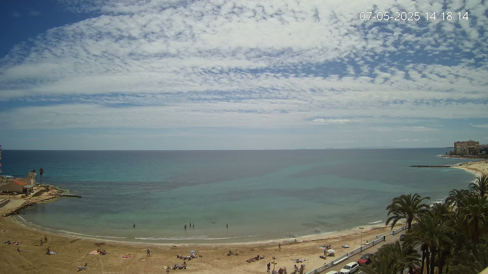 A sandy beach curves around a calm, teal-green sea under a mostly sunny sky with scattered cumulus clouds.