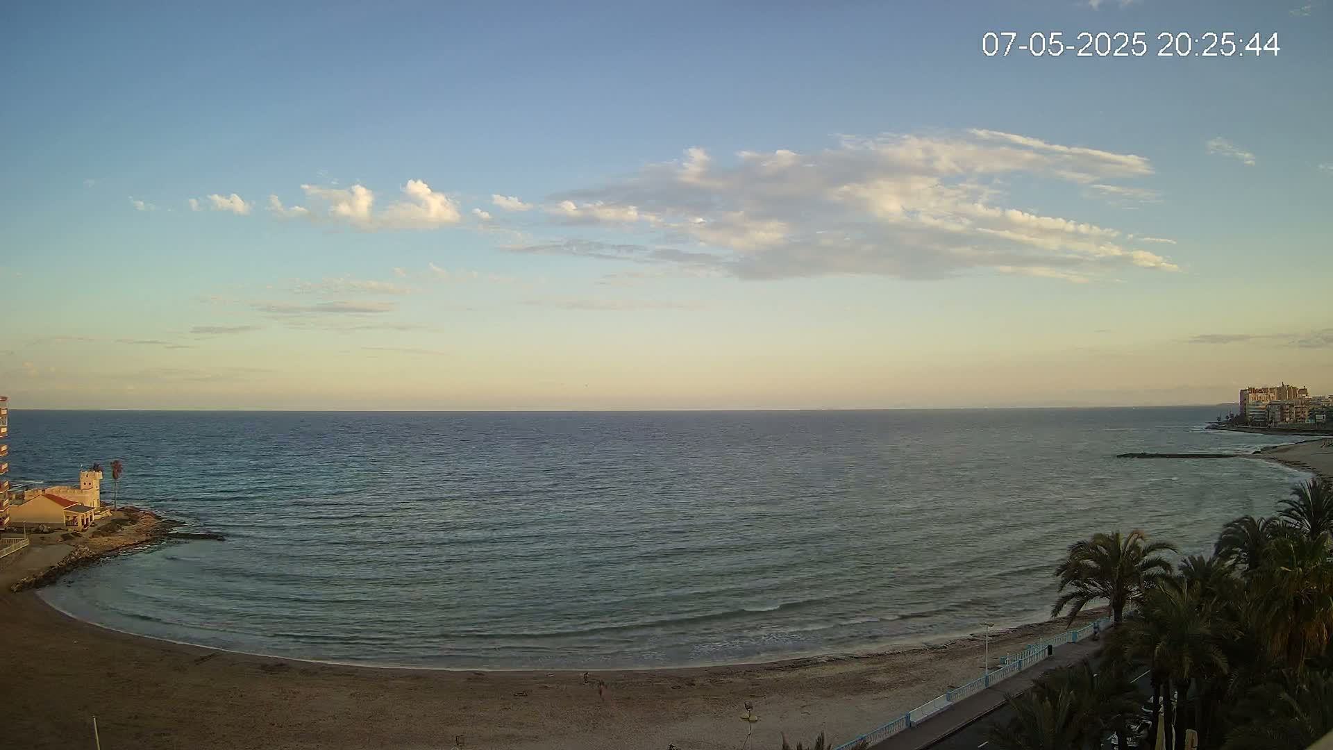 A calm sea with a sandy beach curves gently around a small building near palm trees under a mostly clear sky with some wispy clouds.