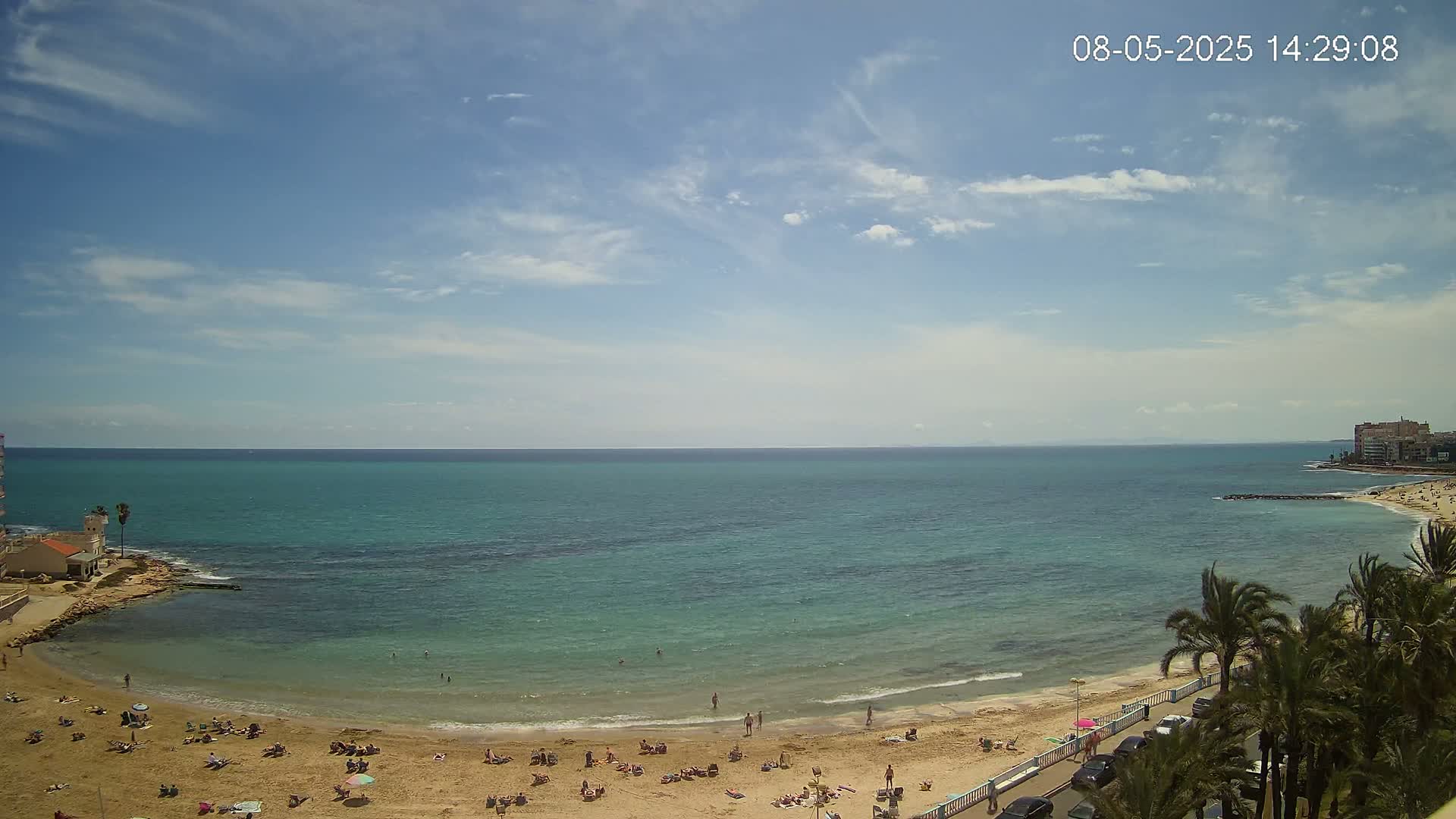 A partly cloudy sunny day shows a sandy beach with scattered people, calm turquoise water, and palm trees lining a road next to the beach.