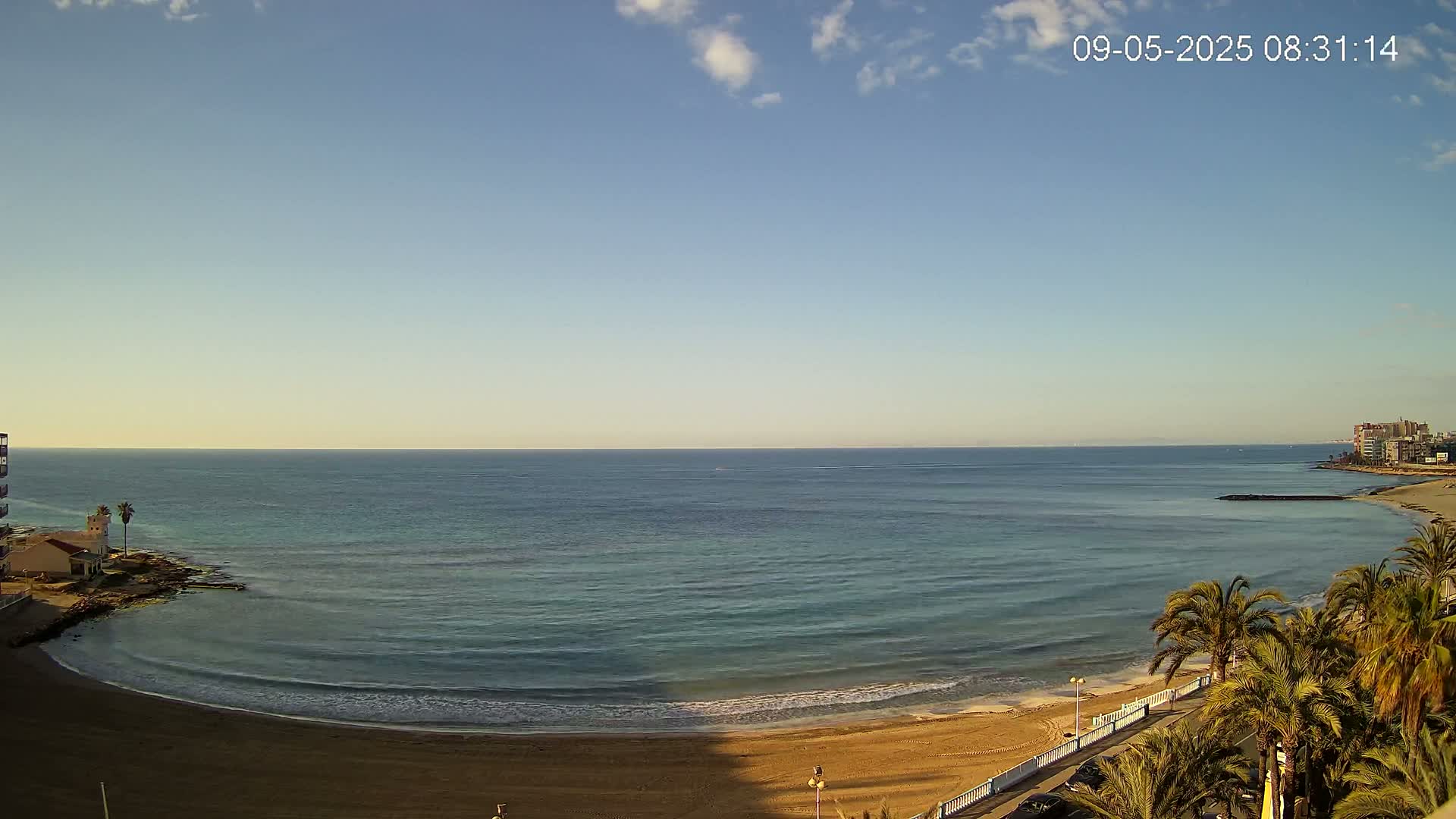 A calm, clear day reveals a sandy beach curving gently into a tranquil teal sea, lined with palm trees and buildings in the distance.