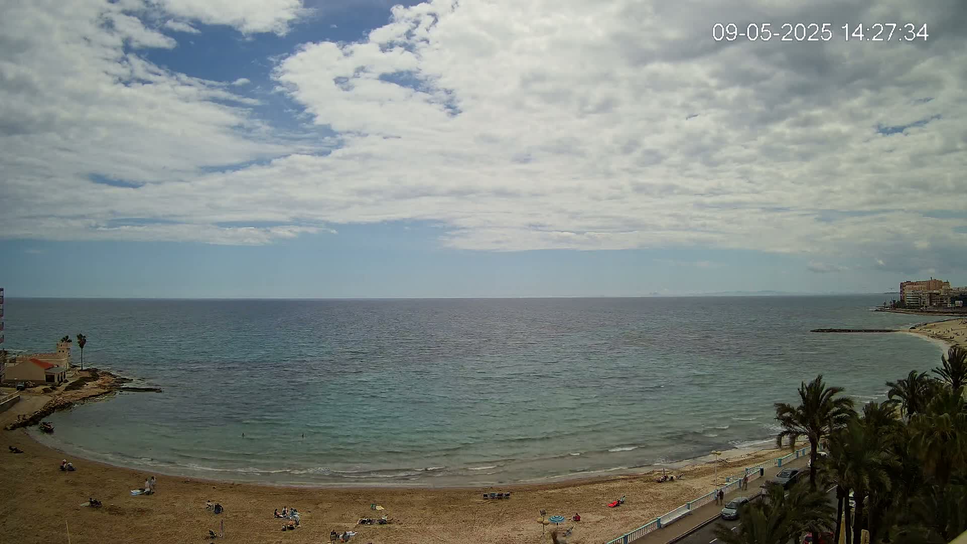 A partly cloudy day reveals a sandy beach curving into a calm, teal sea, with palm trees lining a coastal road and a few people visible on the sand.