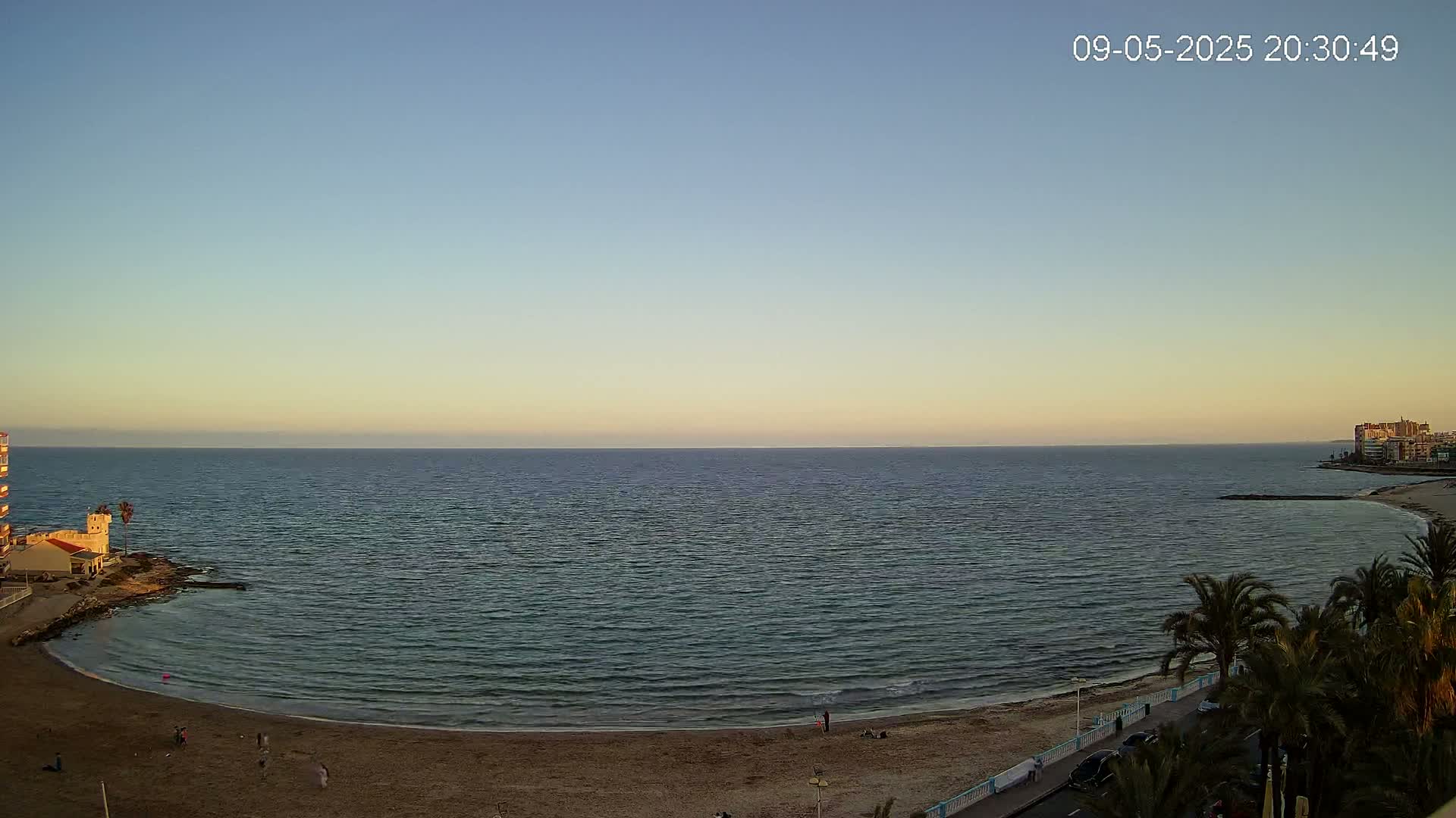 A calm sea laps a sandy beach at sunset, with a few people visible on the shore and palm trees lining a nearby road.