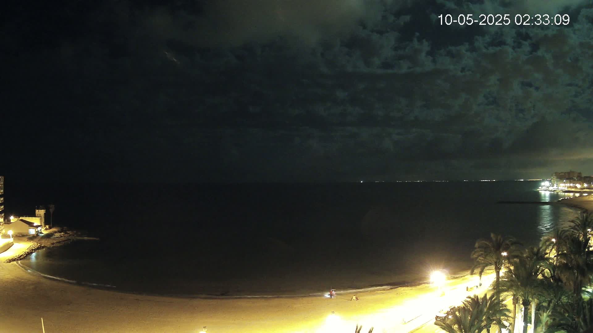 A nighttime view of a dark, cloudy sky over a calm, dark sea with a lit beach and palm trees along a promenade.