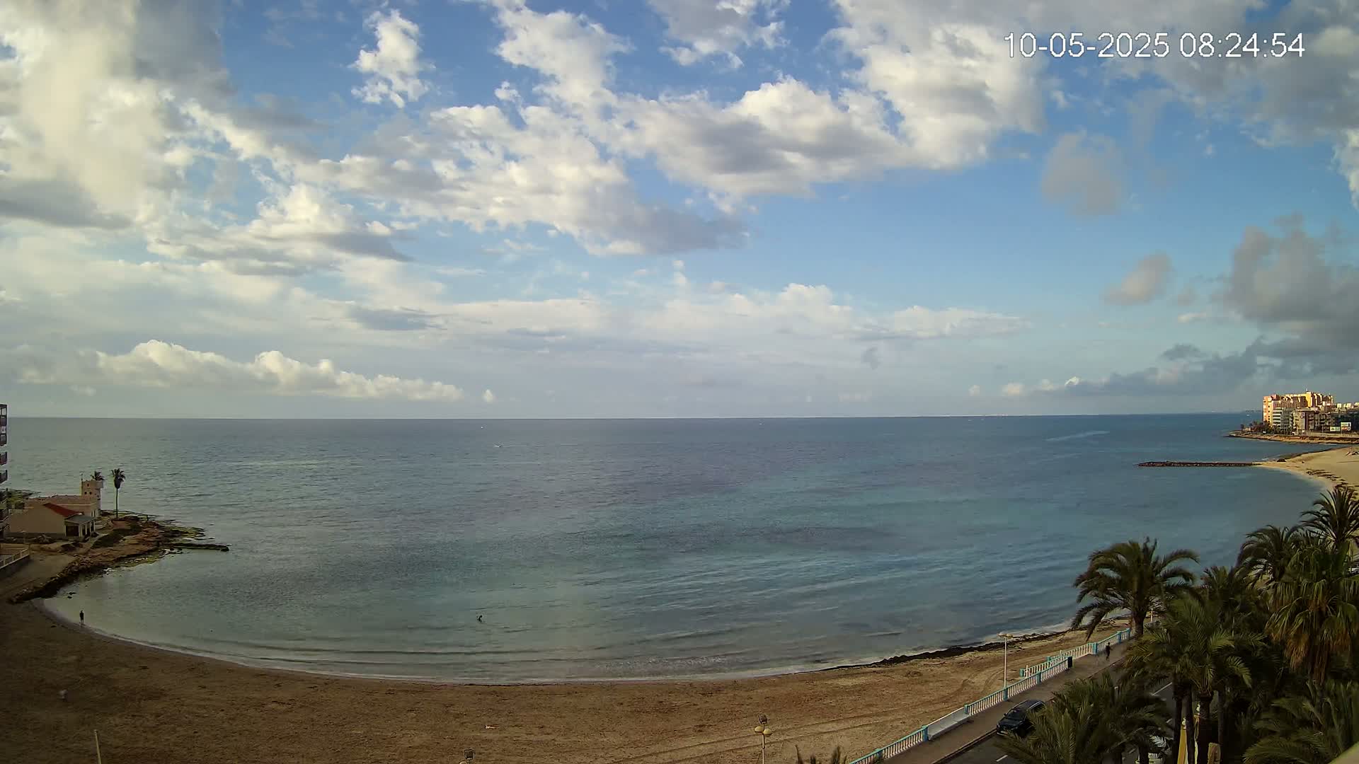 A partly cloudy sky overlooks a calm, blue sea curving around a sandy beach lined with palm trees and buildings.