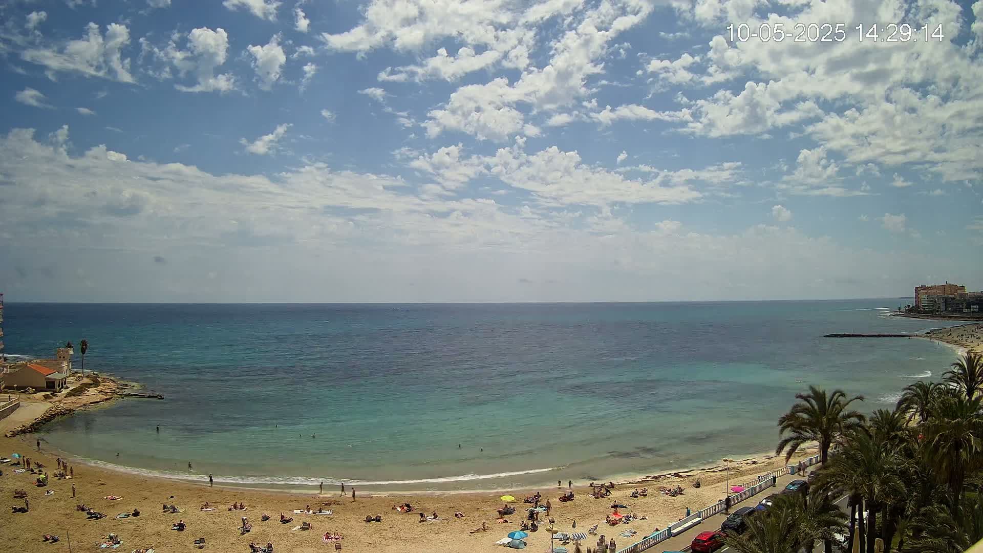 A sandy beach with scattered sunbathers curves around a calm, teal-colored bay under a partly cloudy sky.