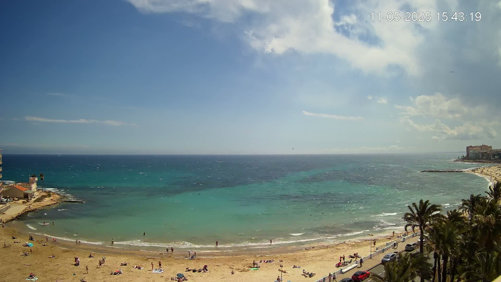 A partly sunny day with a windy ocean shows a sandy beach with scattered sunbathers, a road with parked cars, and palm trees lining the shore.