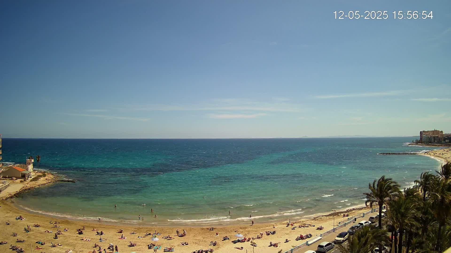 A sunny day shows a beach with many people relaxing on the sand, palm trees lining a road nearby, and a calm, teal ocean stretching to a distant coastline.