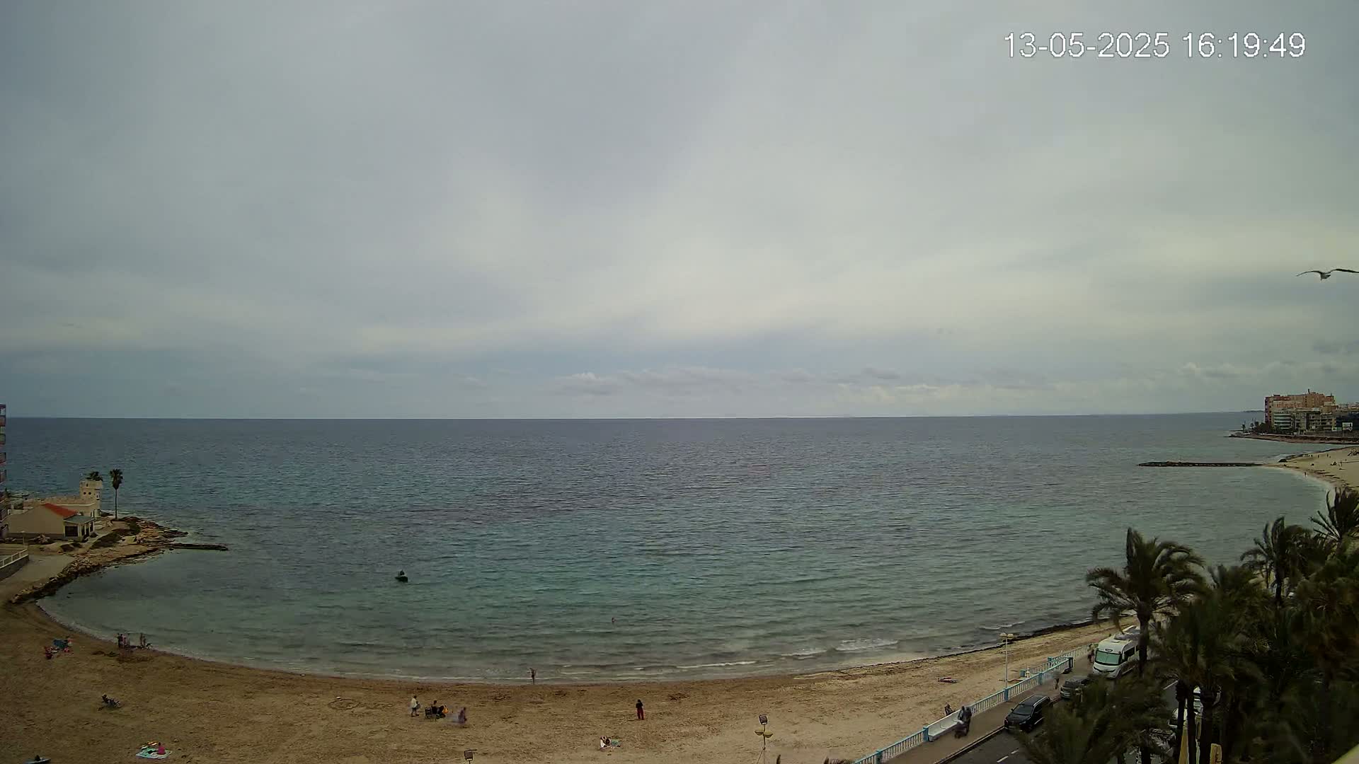 A sandy beach curves around a calm, teal sea under an overcast sky, with a few people scattered on the beach and palm trees lining the road beyond.