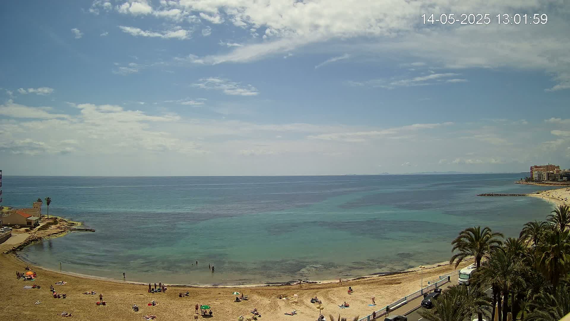A sandy beach with scattered people curves around a calm, clear teal ocean under a partly cloudy sky.