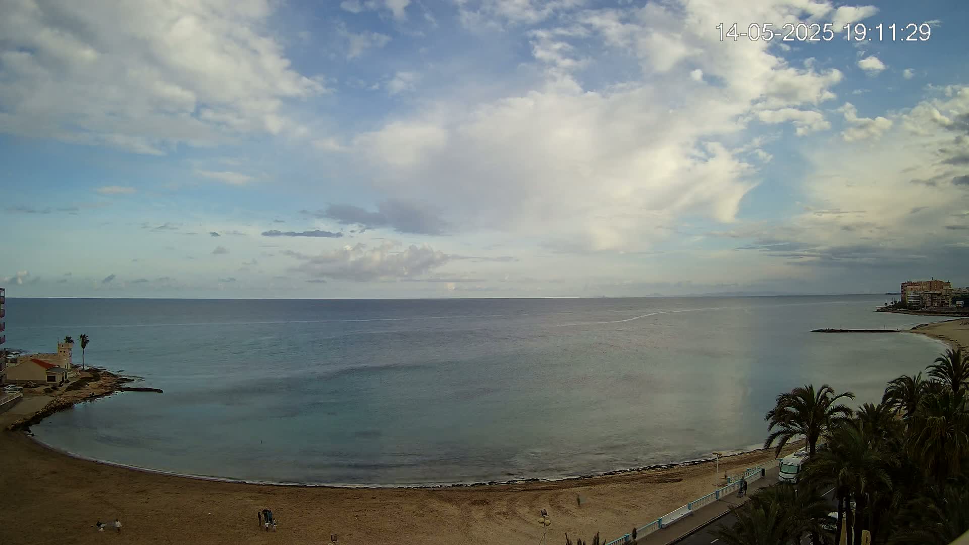 A partly cloudy sky overlooks a calm, teal ocean curving around a sandy beach with palm trees and a few buildings visible in the distance.