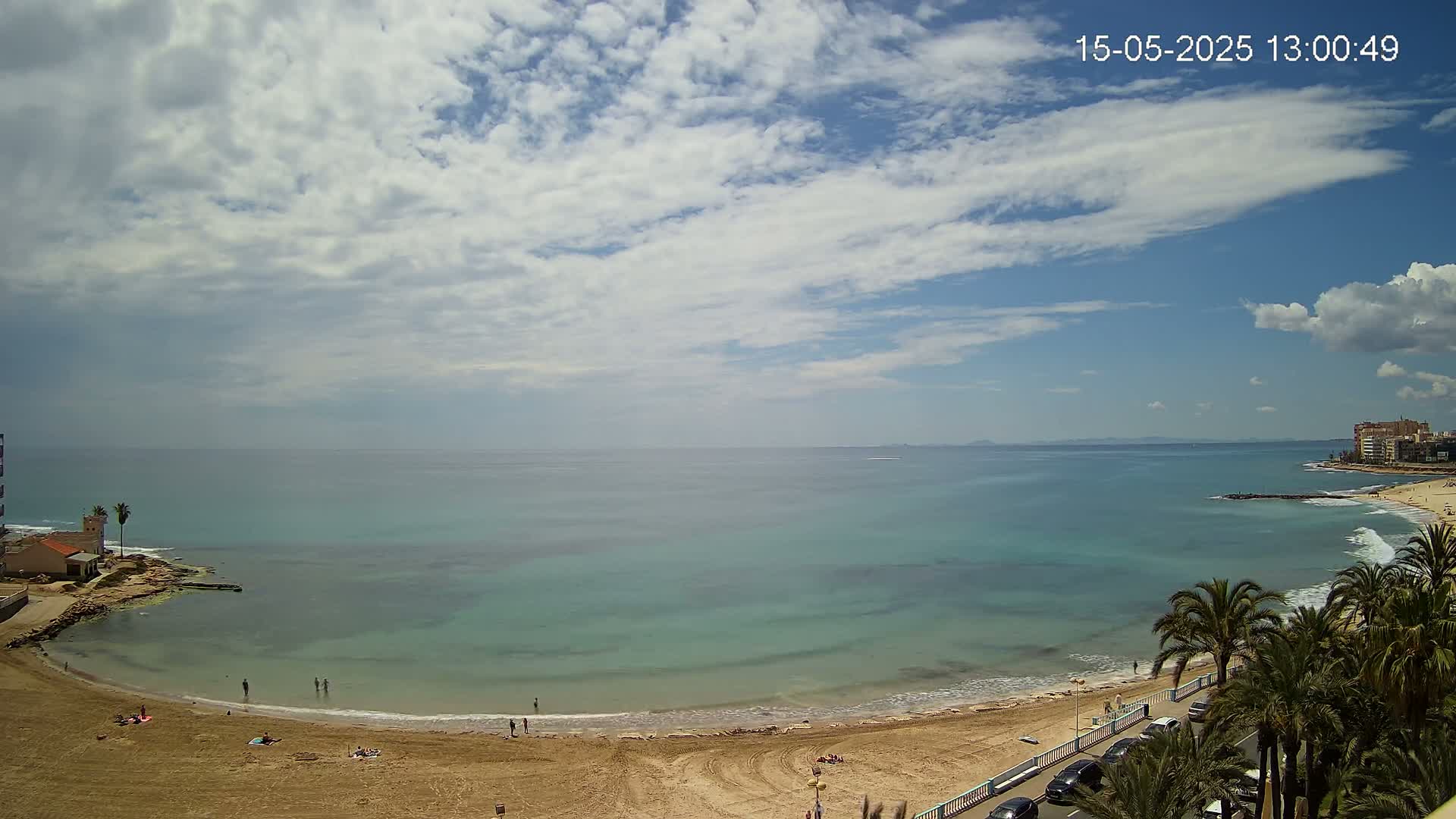 A partly cloudy sky overlooks a calm, teal ocean meeting a sandy beach with a few people scattered across it, palm trees lining the road beyond.