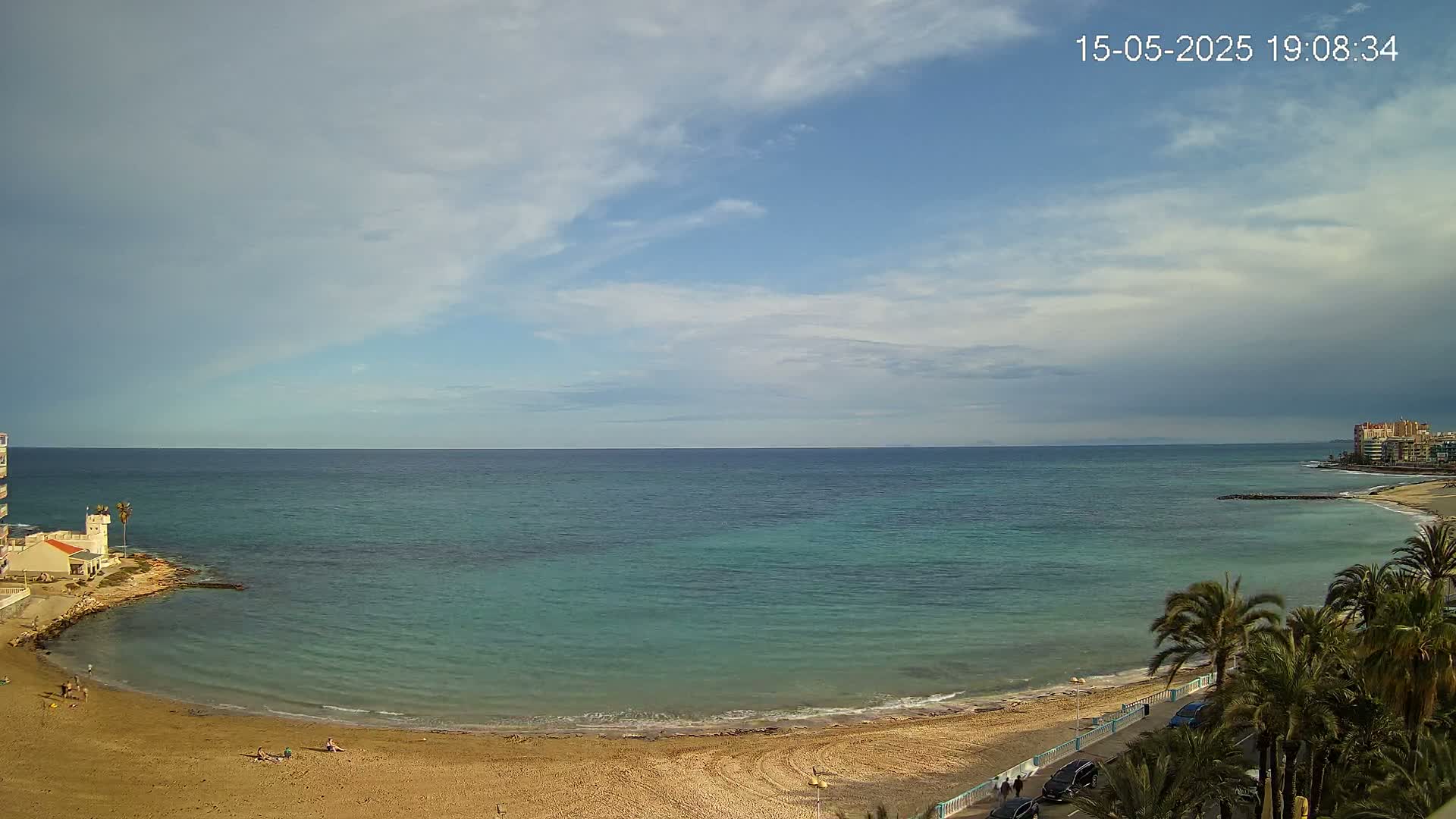 A sandy beach curves around a calm, teal ocean under a partly cloudy sky.