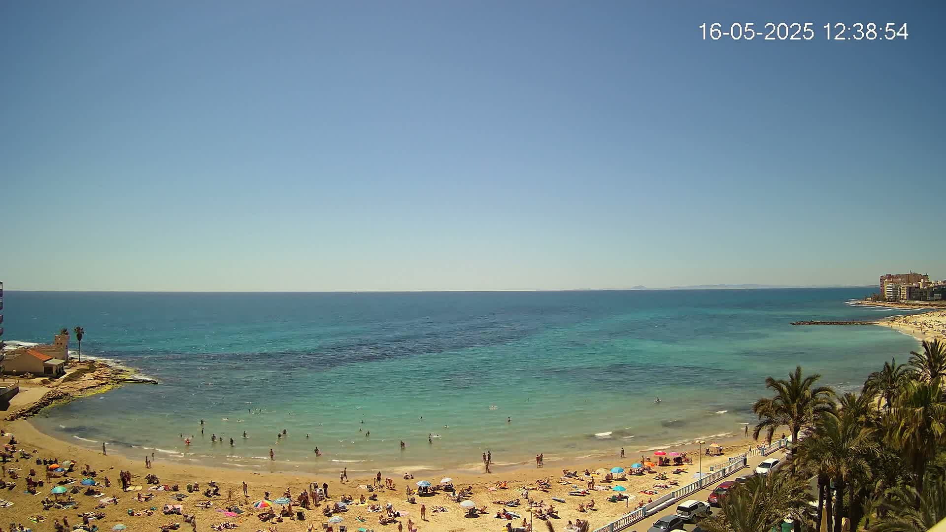 A sunny day at a beach shows calm, clear turquoise water lapping a sandy shore crowded with sunbathers and palm trees lining the road beyond.
