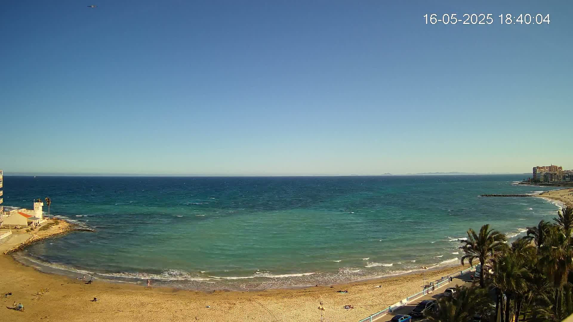 A sandy beach with a few people on it curves around a calm, teal-colored bay that opens to a blue ocean under a clear blue sky.